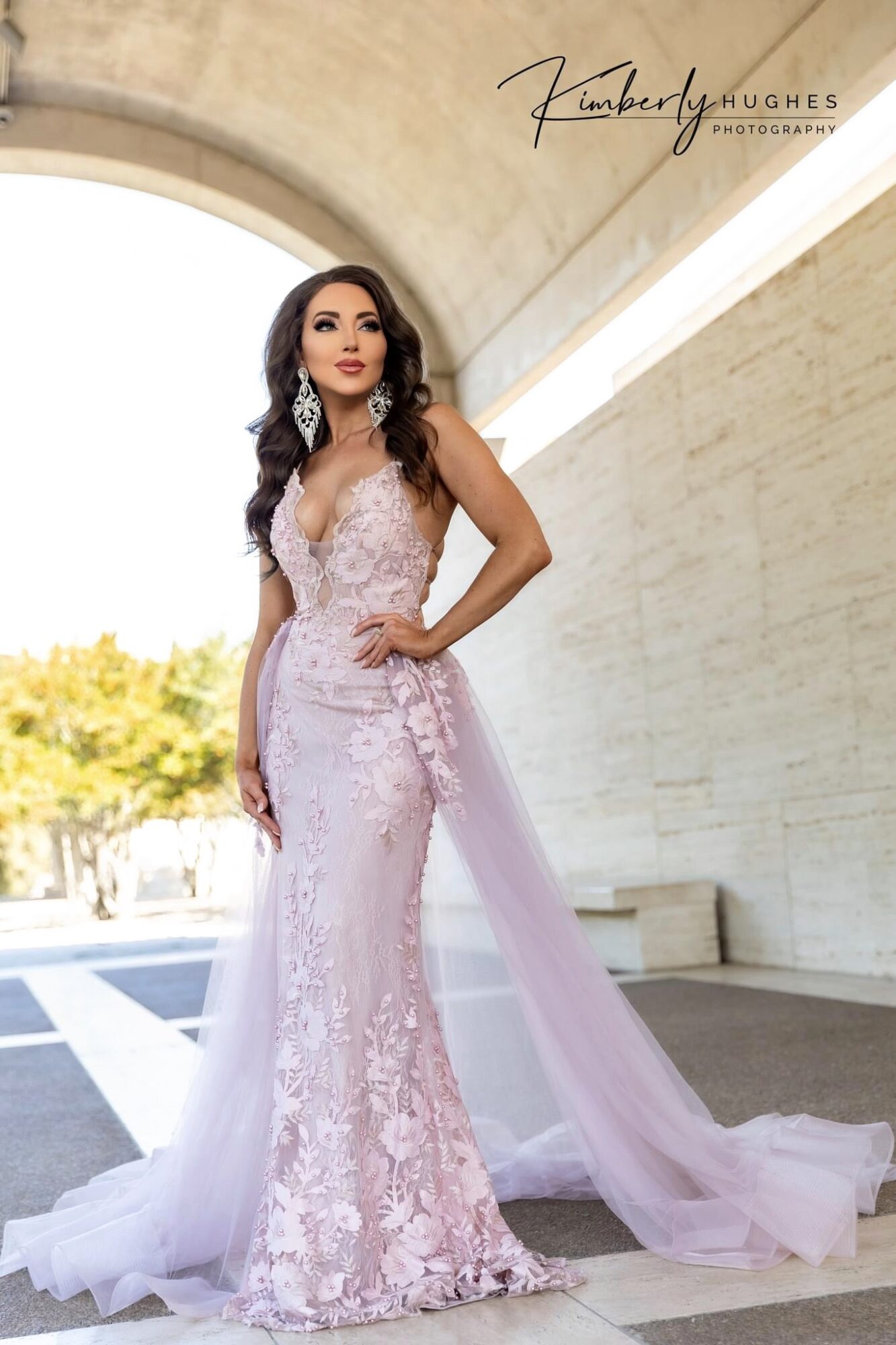 Woman in a pink lace gown with a train, standing under an archway with trees in the background.