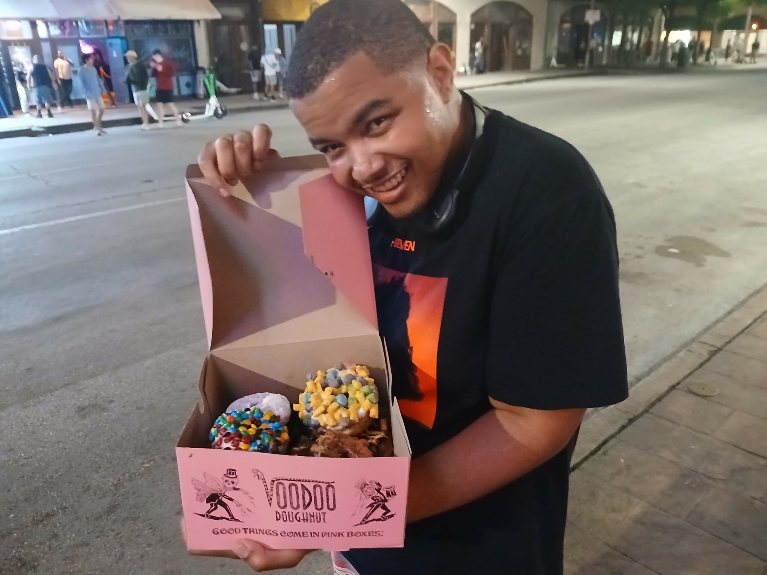 Boy holding a box of decorated cupcakes outdoors at night, smiling, with people in the background.