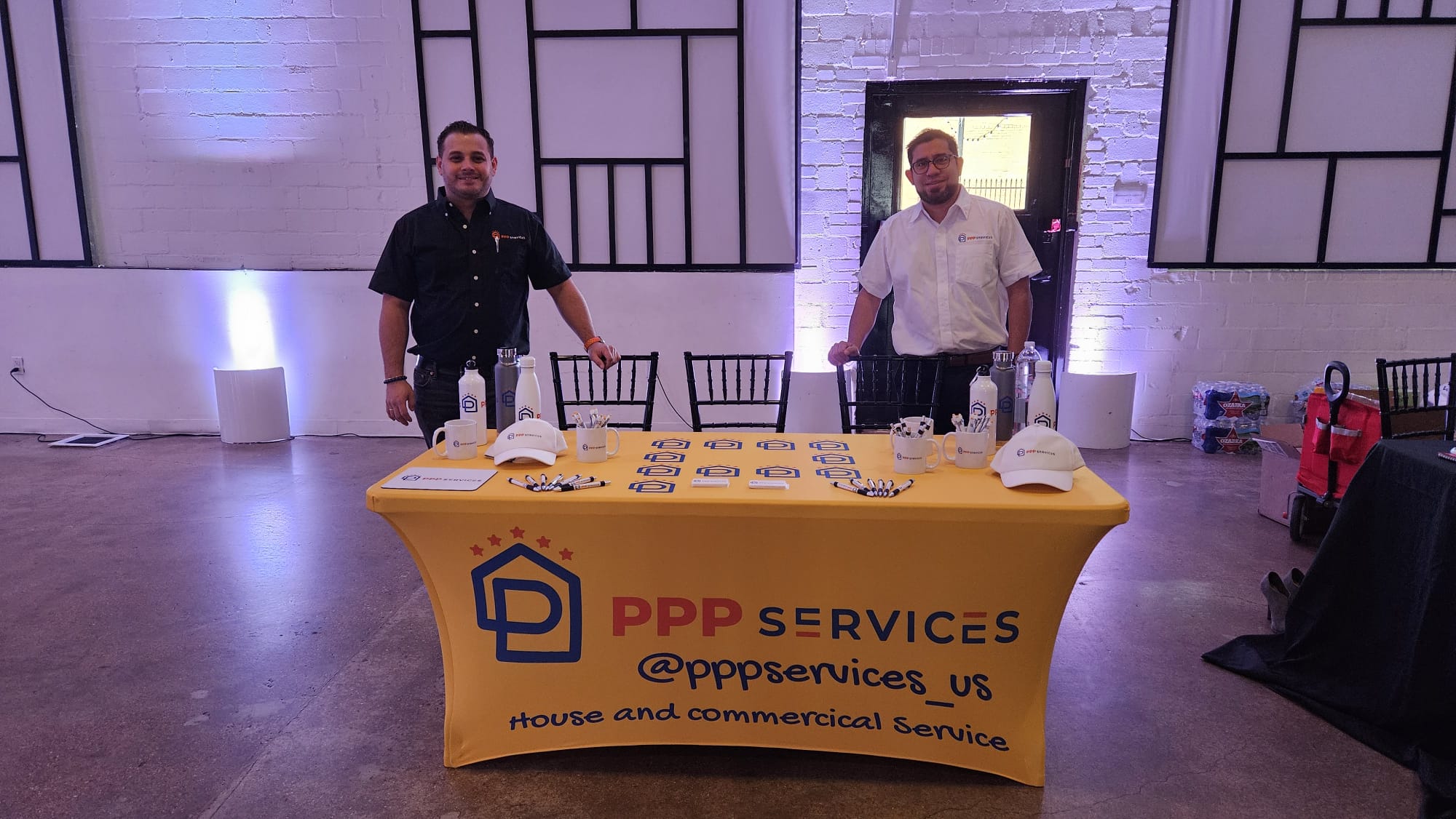 Two men standing behind a table with a yellow tablecloth displaying 'PPP SERVICES' and contact information, in a room with modern decor.