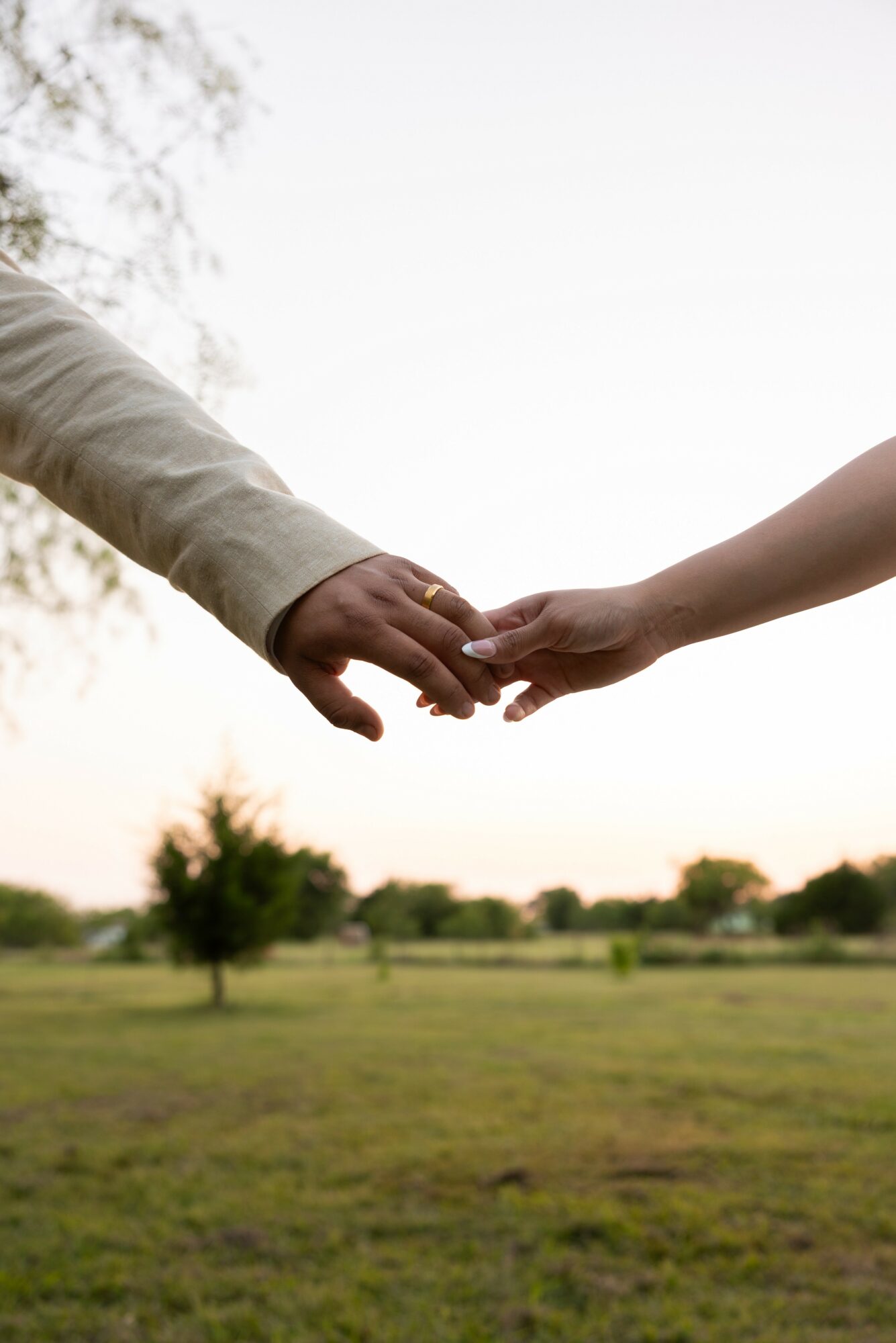 Two people reaching out and holding hands outdoors with trees and grass in the background.