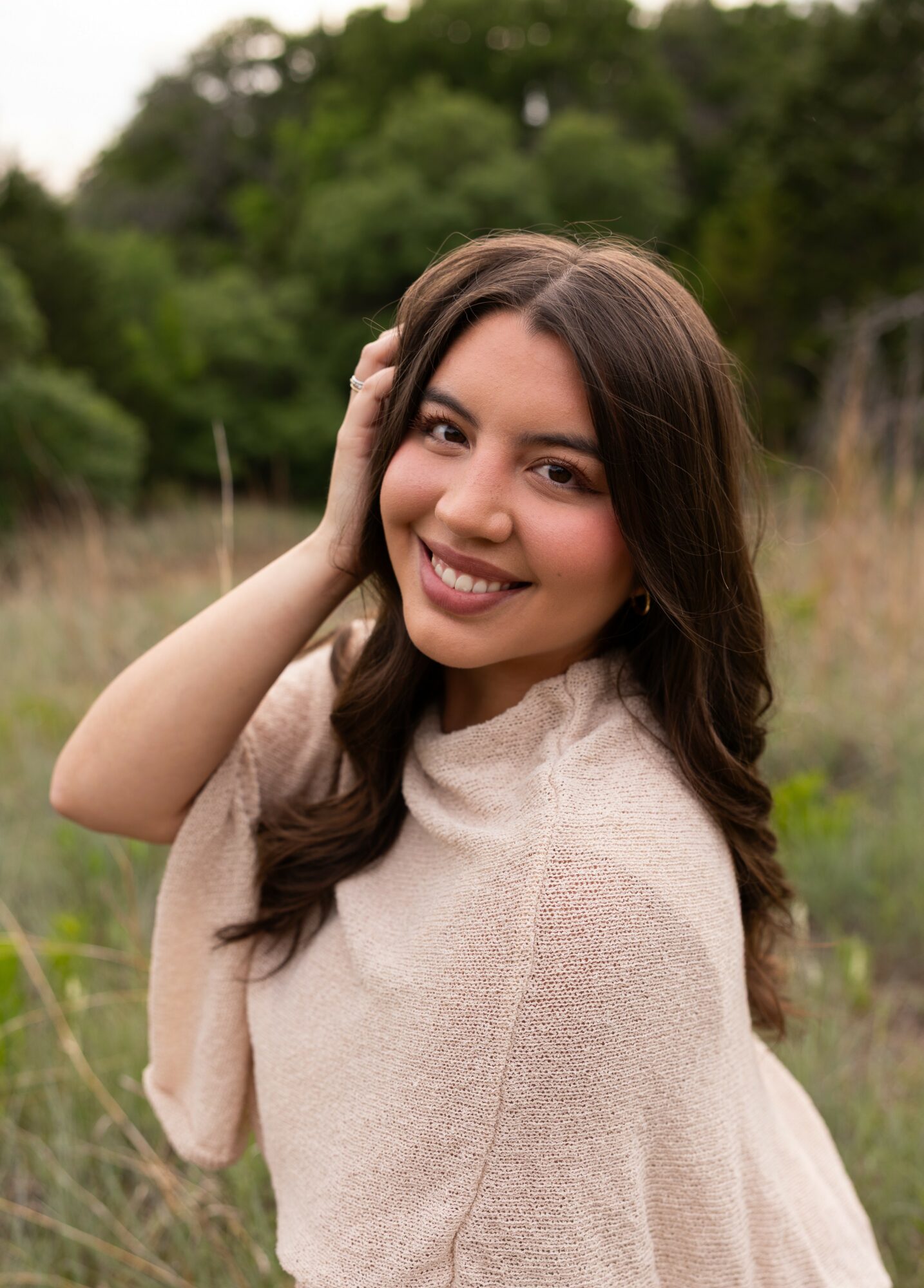Young woman with long dark hair smiling outdoors, hand on her head, wearing a light-colored top, greenery in background.