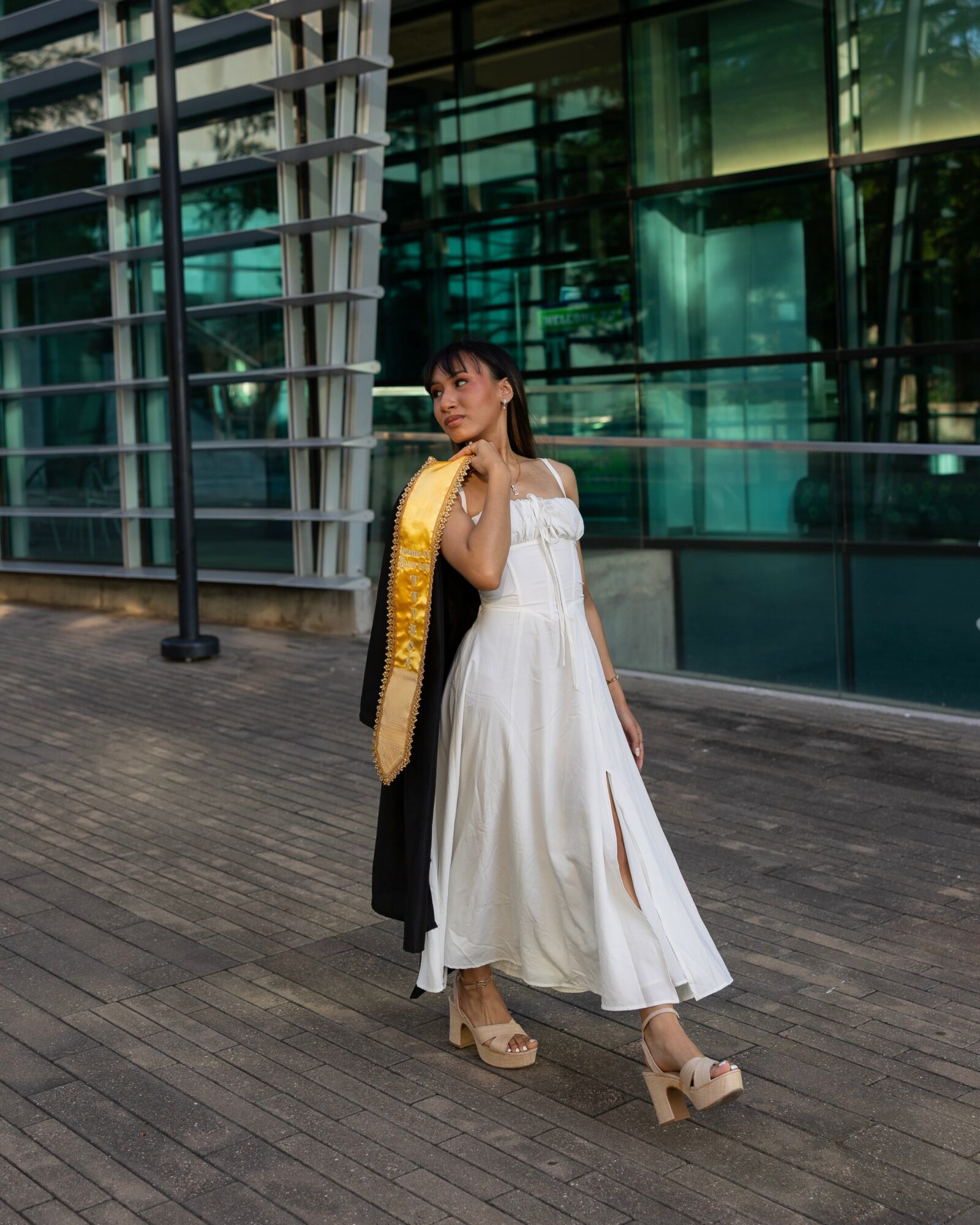 Woman in white dress and beige heels standing outside near modern glass building with metal structures.