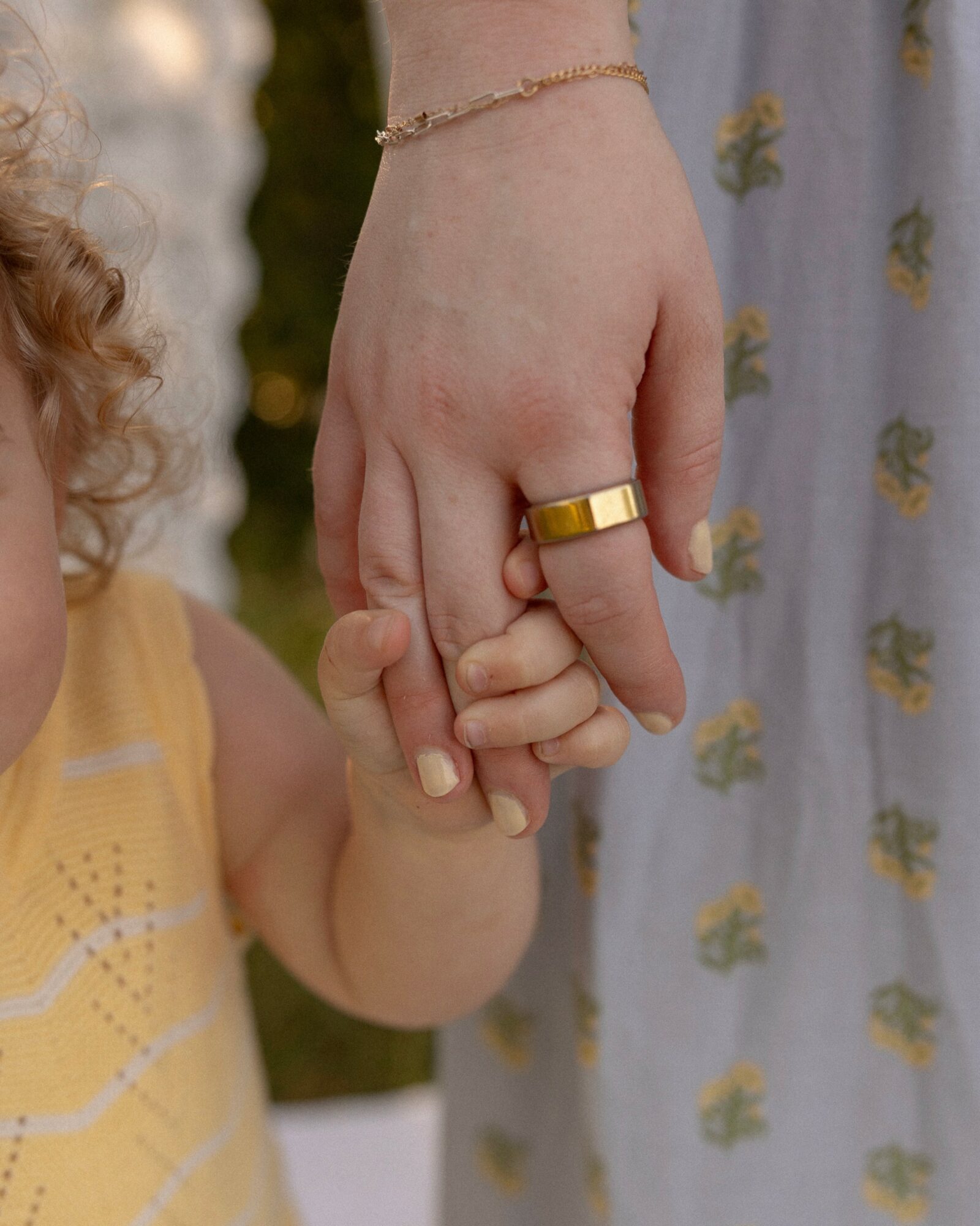 Close-up of an adult and child holding hands, adult wearing a gold ring and bracelet, child in yellow clothing.