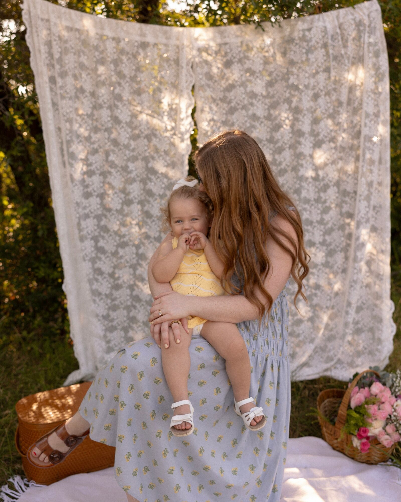 Woman holding a smiling child outdoors with a lace backdrop, flowers in a basket nearby, sunlight filtering through trees.