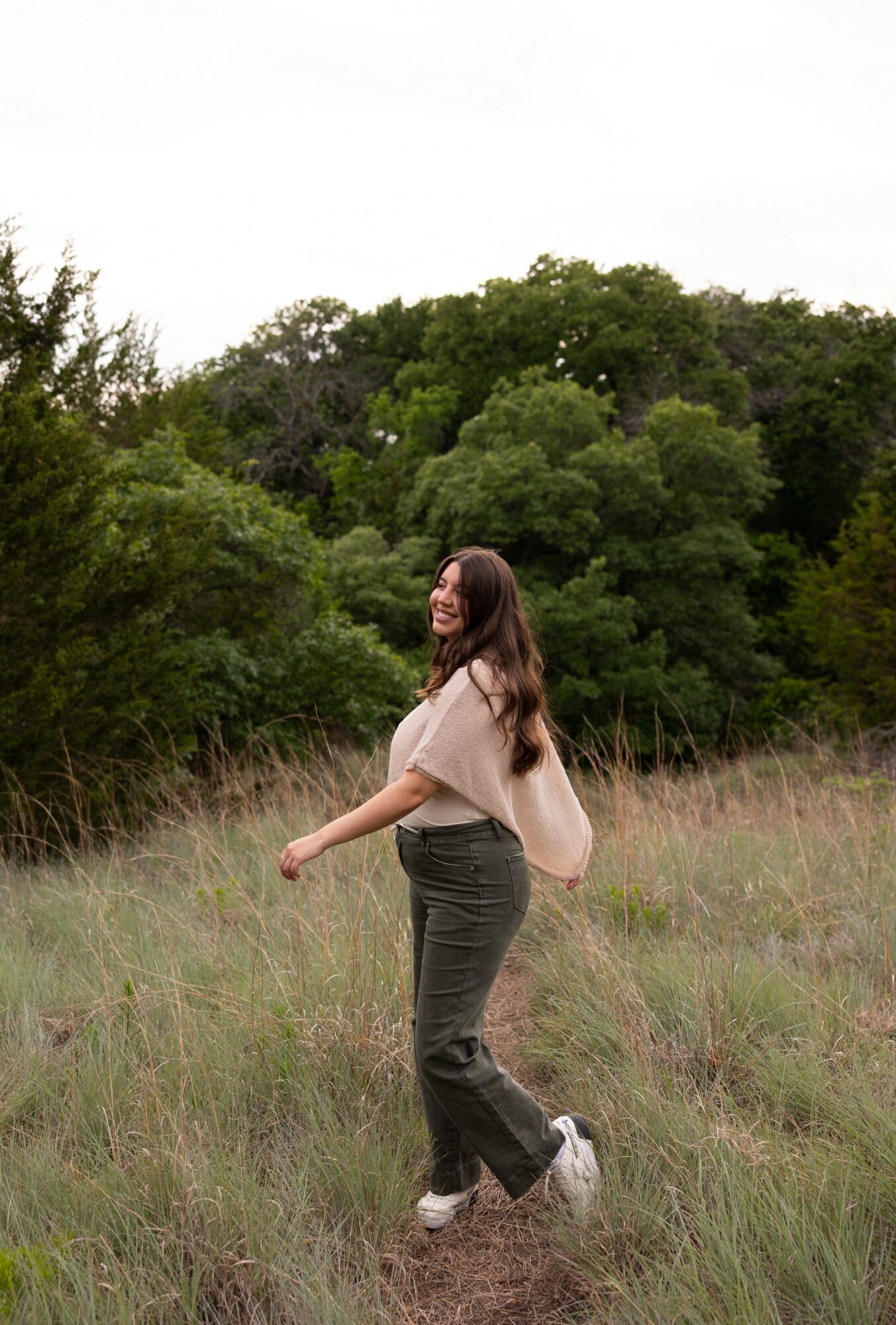 Young woman walking through grassy field with trees and bushes in background, smiling, wearing a light top and dark pants.