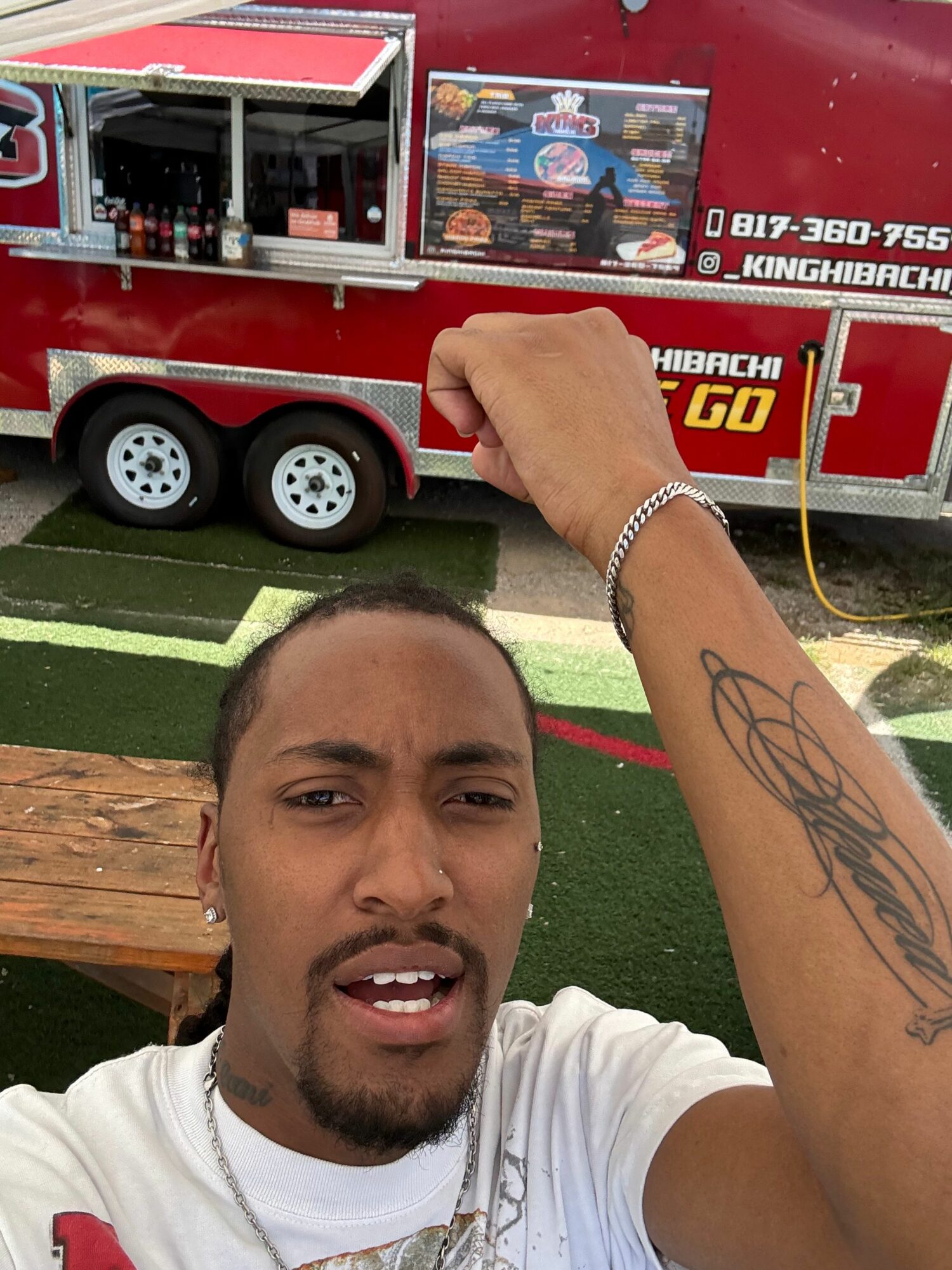 Young man with a tattoo on his arm taking a selfie, smiling, with a red food truck in the background.