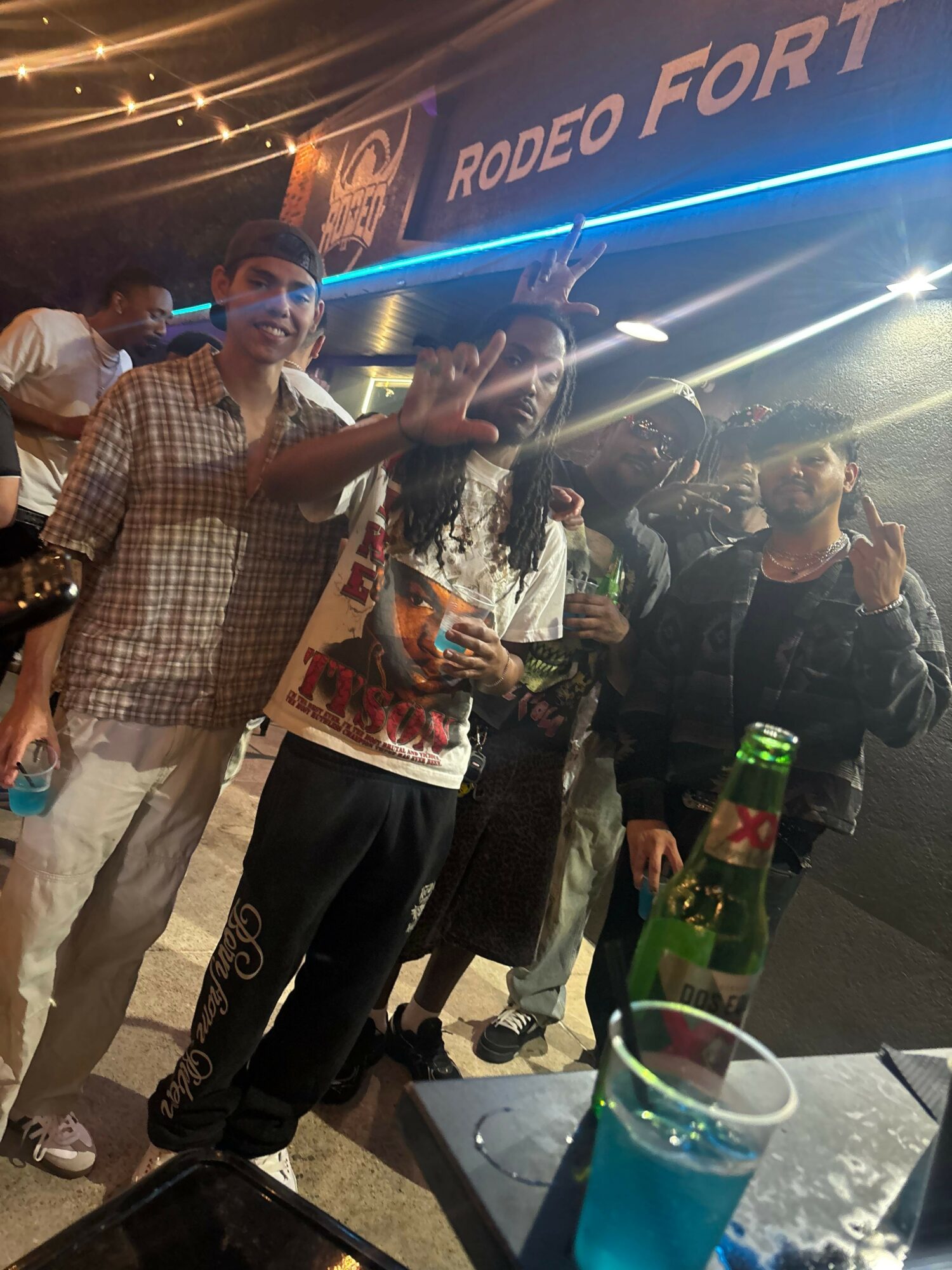 Group of people standing in front of a neon sign that reads 'RODEO FORA', with drinks on a table nearby.
