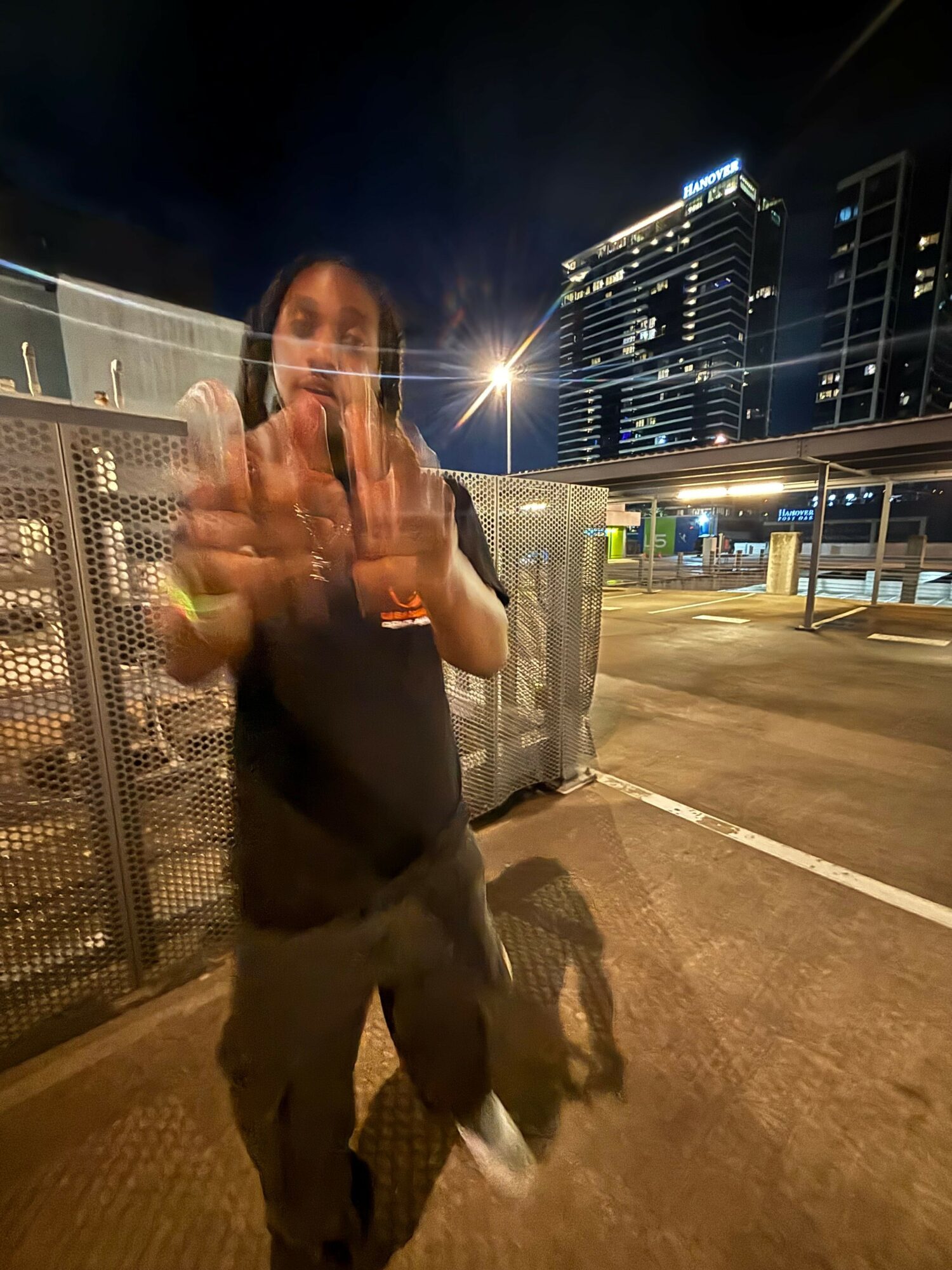 Person standing outdoors at night, pointing towards the camera, with city buildings and bright lights in the background.