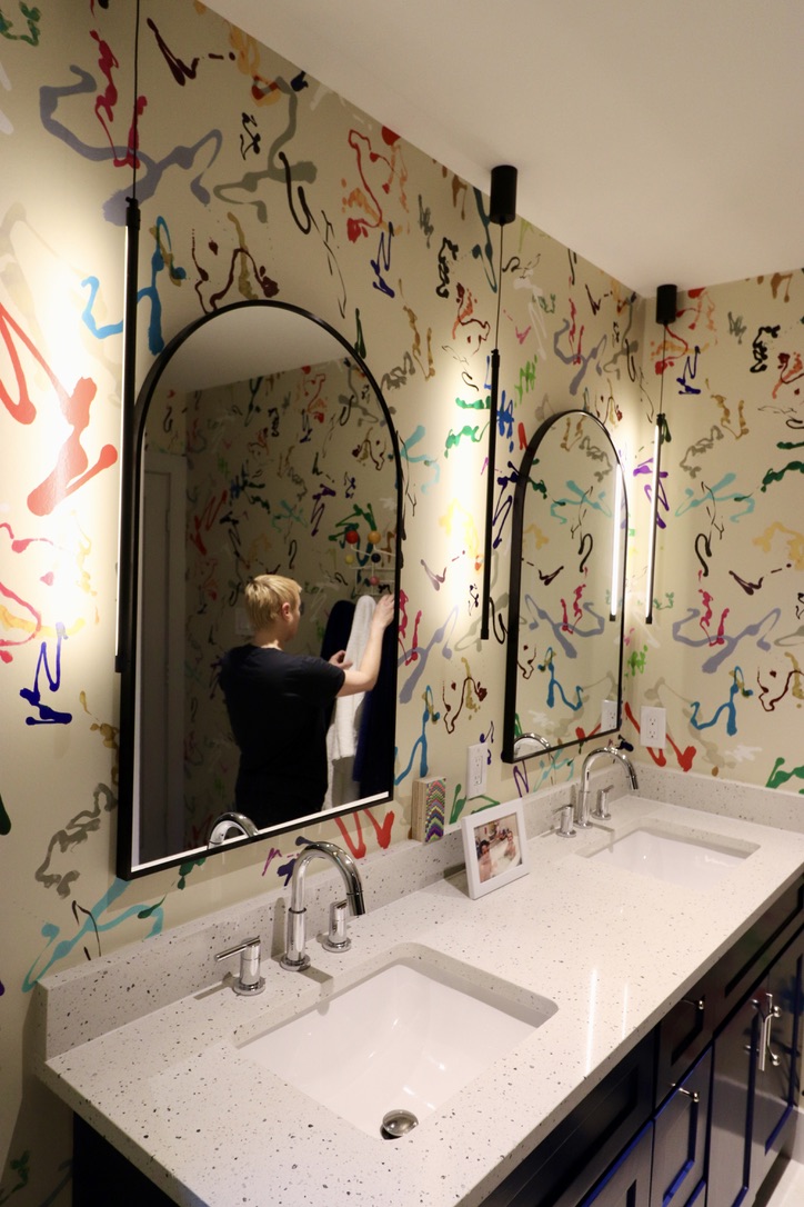 Bathroom with two mirrors above a white countertop with two sinks, colorful abstract wall art, and a person taking a photo in the mirror.