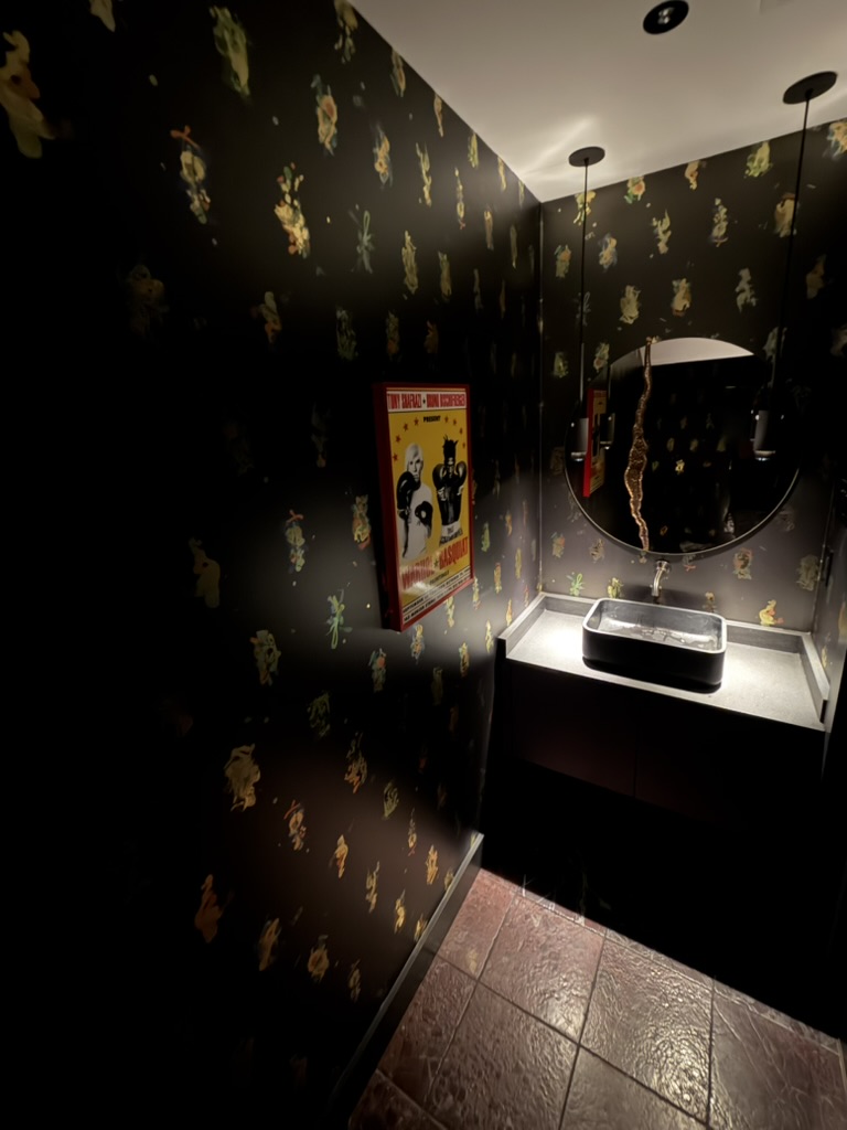 Bathroom with black wallpaper, round mirror, illuminated sink, and a colorful poster on the wall.
