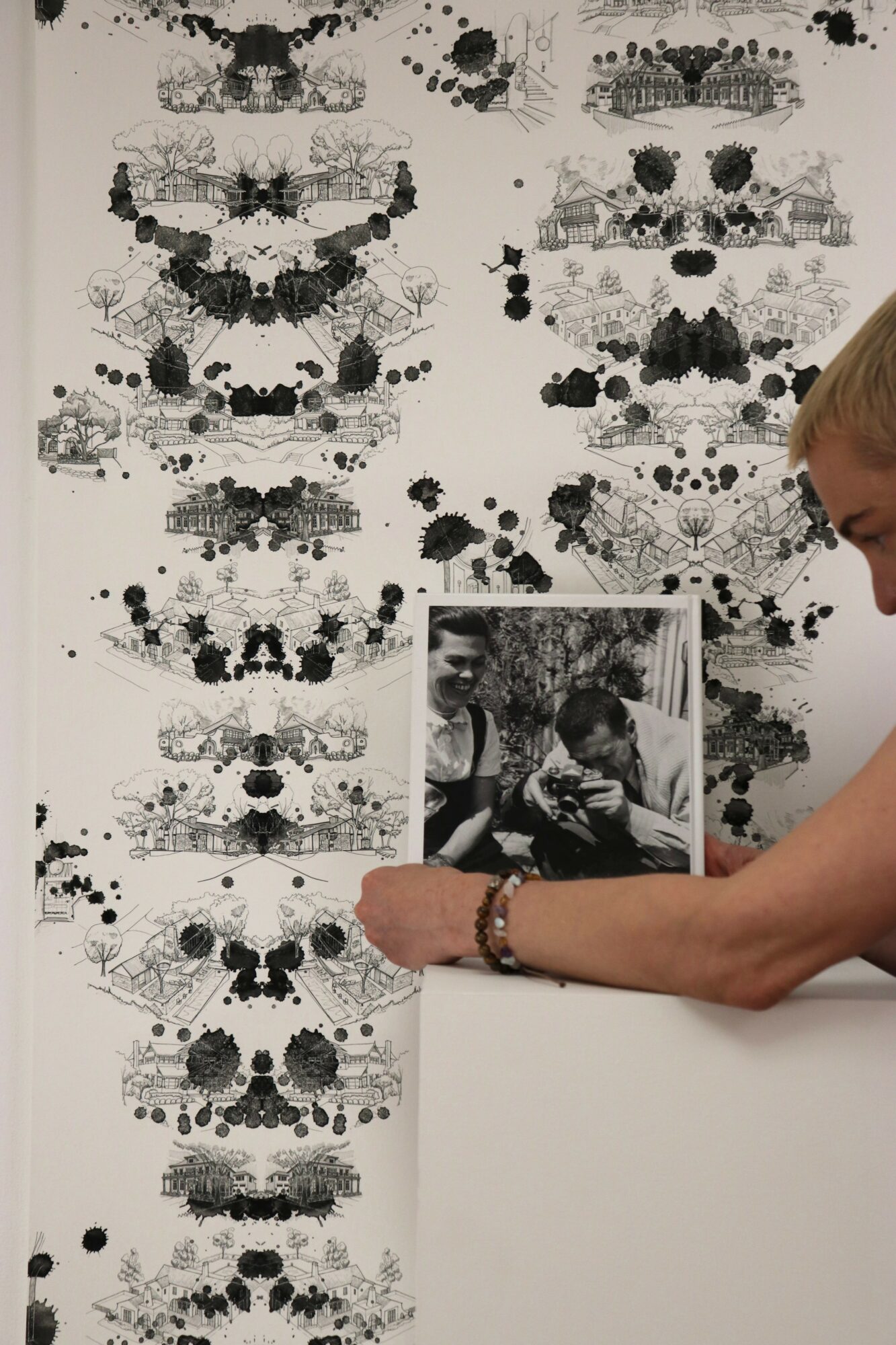 Person holding a black-and-white photo in front of a large abstract black inkblot artwork on a white wall.