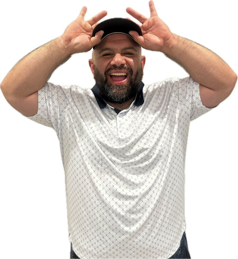 Man with beard smiling, wearing a white patterned shirt and black cap, making playful hand gestures near his head.