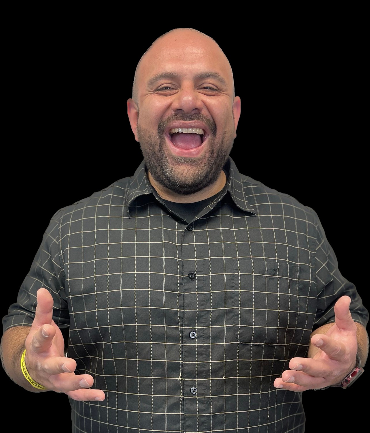 A smiling man with a beard and bald head, wearing a checkered shirt, gestures with both hands, against a black background.