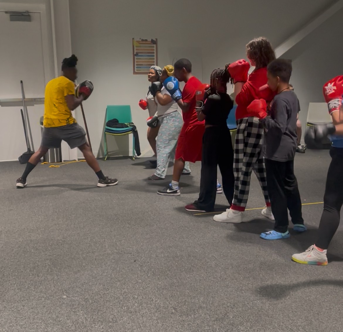 Children and adults in a boxing class, some wearing boxing gloves, in a room with a gray carpeted floor.