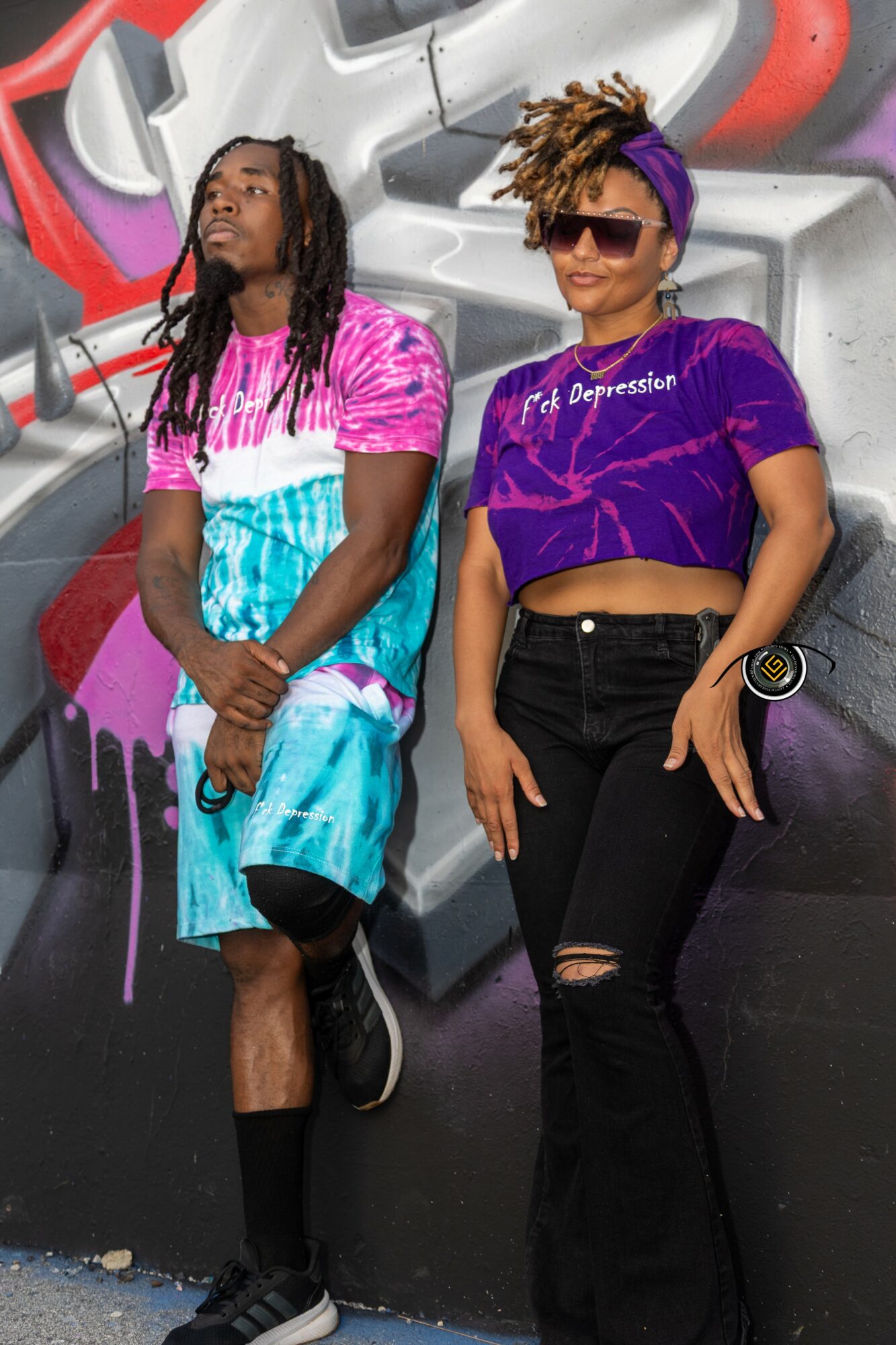 Two women stand in front of a graffiti-covered wall, one wearing a pink and blue outfit, the other in a purple top and black pants, both with sunglasses.