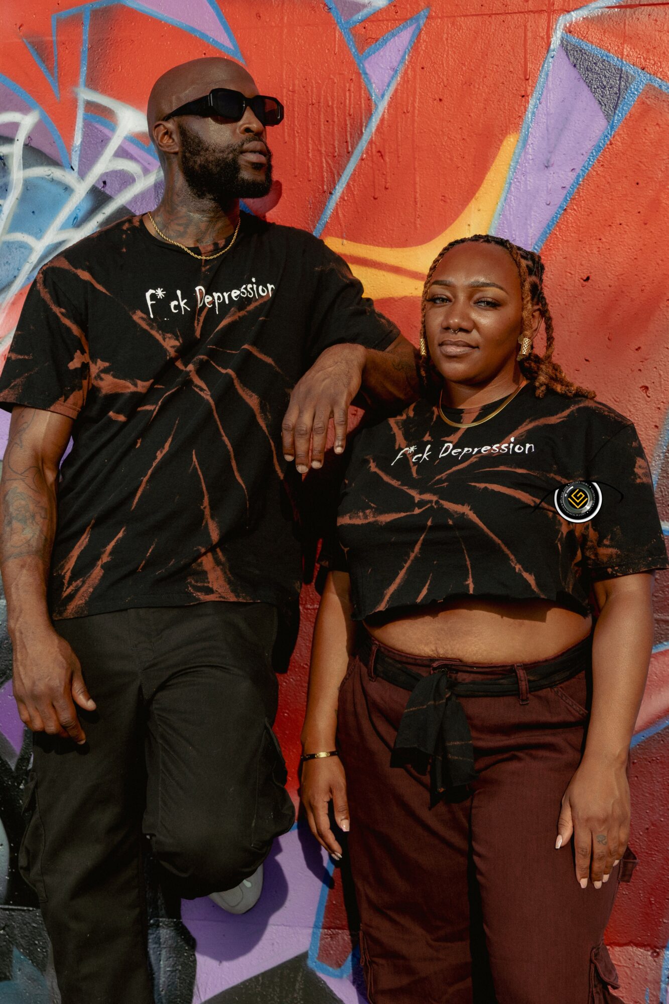 Two people stand in front of a colorful graffiti wall, wearing matching black shirts with white text, one with sunglasses.
