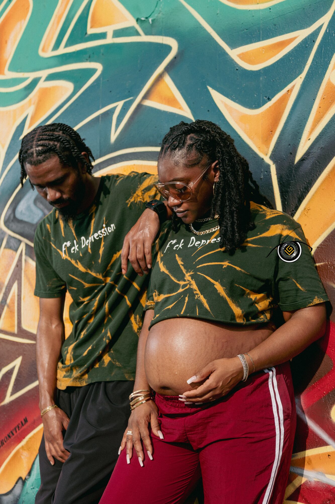 A pregnant woman and a man stand in front of a colorful graffiti wall, both wearing matching green and yellow shirts.