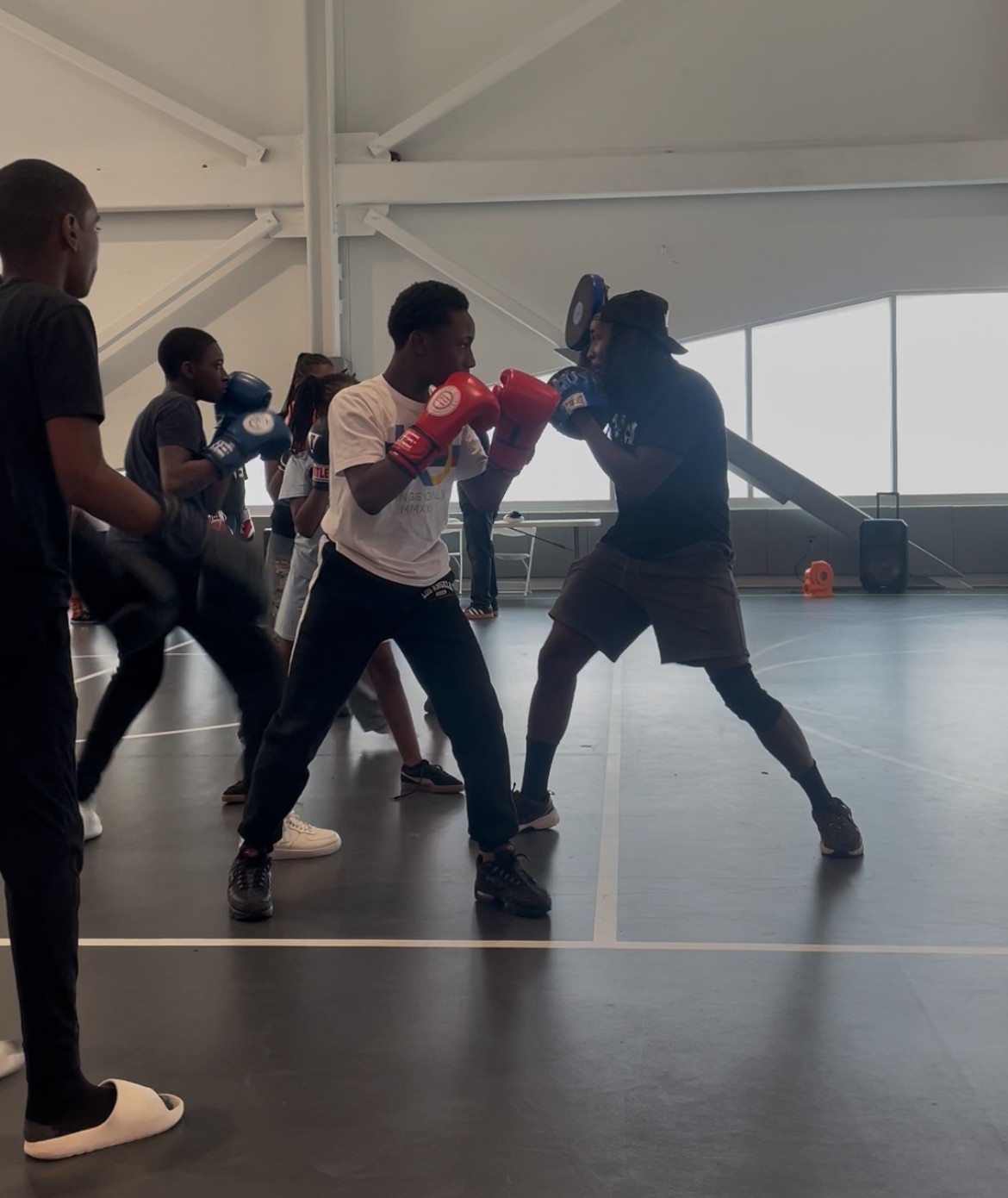 Group of people practicing boxing in a gym, with two individuals sparring and others observing, inside a spacious indoor facility.