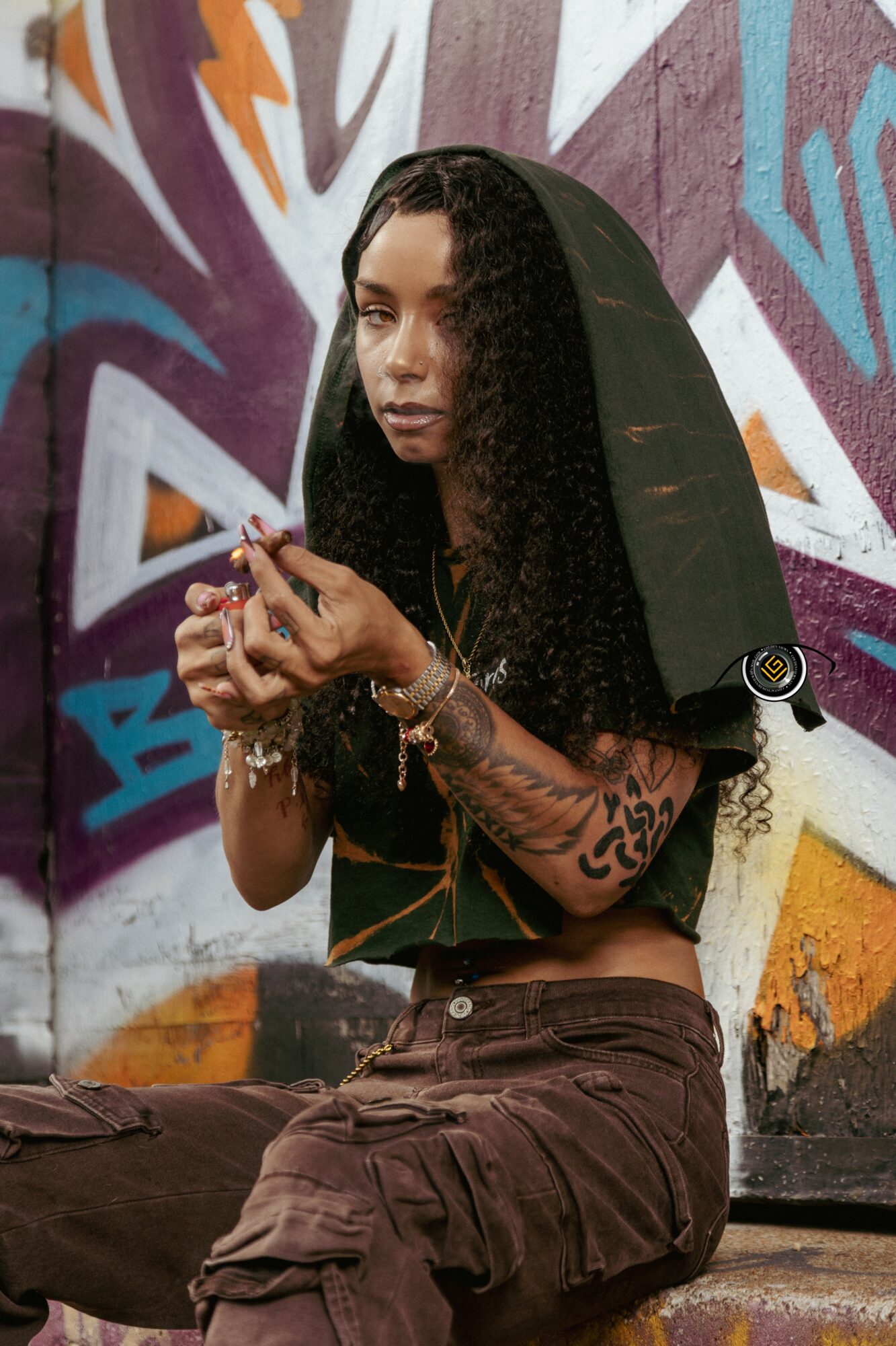 Young woman with curly hair and tattoos, wearing a black hoodie, sitting against a colorful graffiti wall, holding a phone.