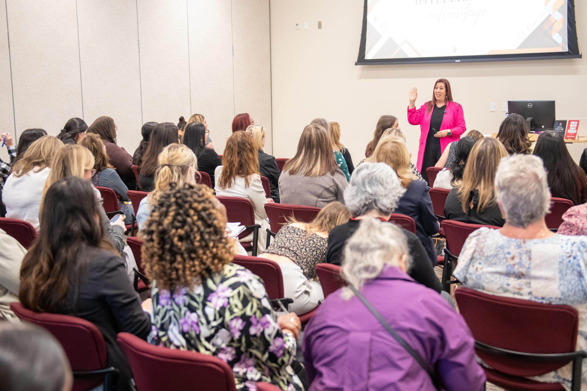 Woman in pink blazer giving a presentation to a seated audience in a conference room.