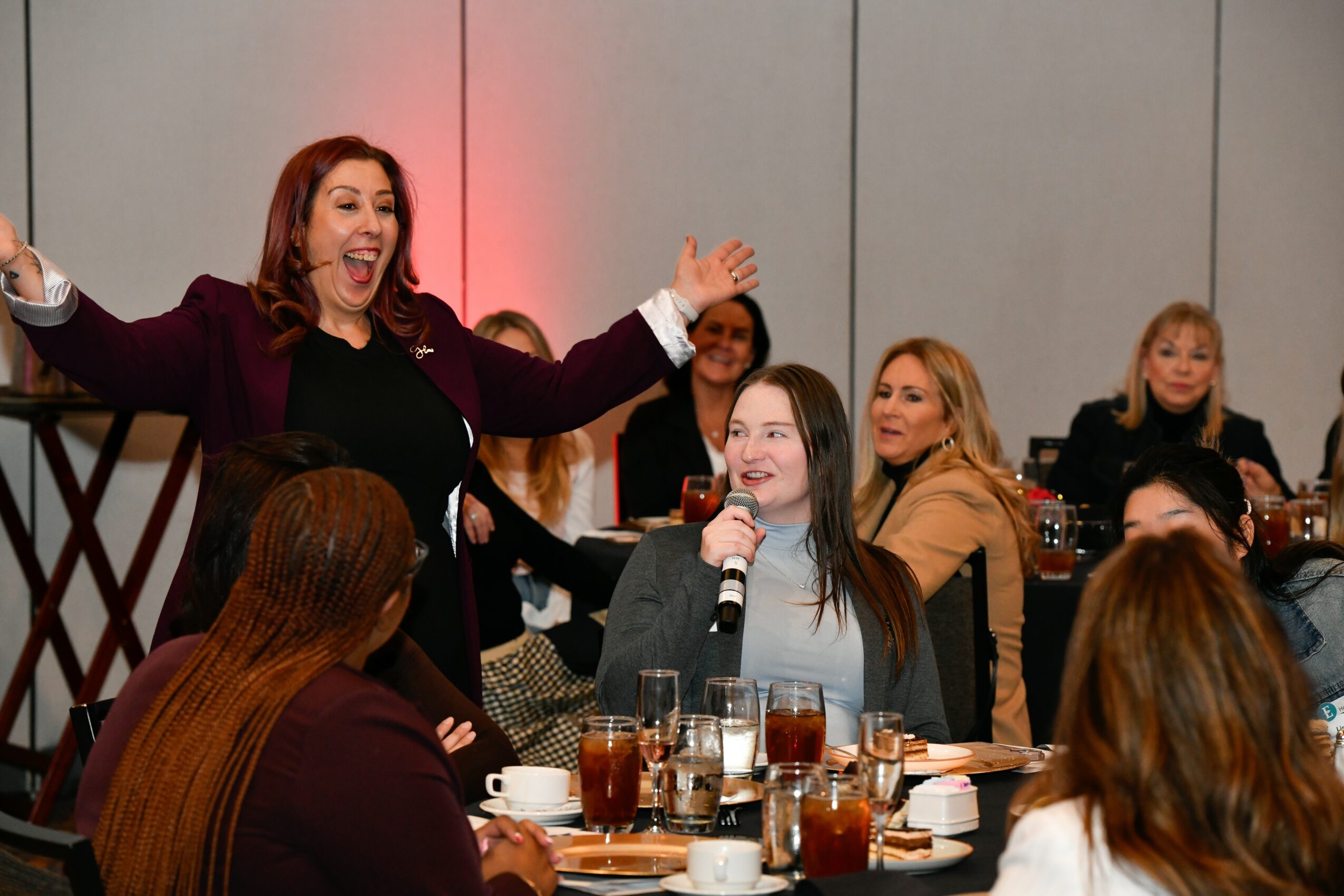 Woman with red hair speaking and gesturing to a group of women seated at a table, smiling and listening, in a social setting.
