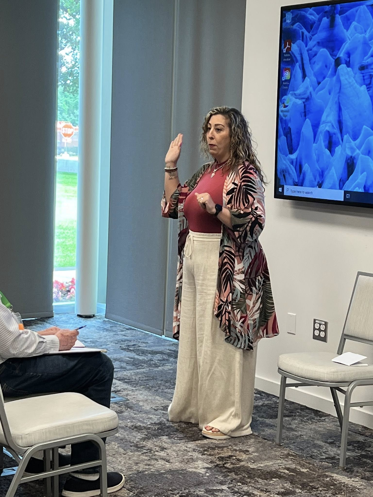 Woman standing and speaking in a room with a large screen on the wall, a chair, and a window with vertical blinds.