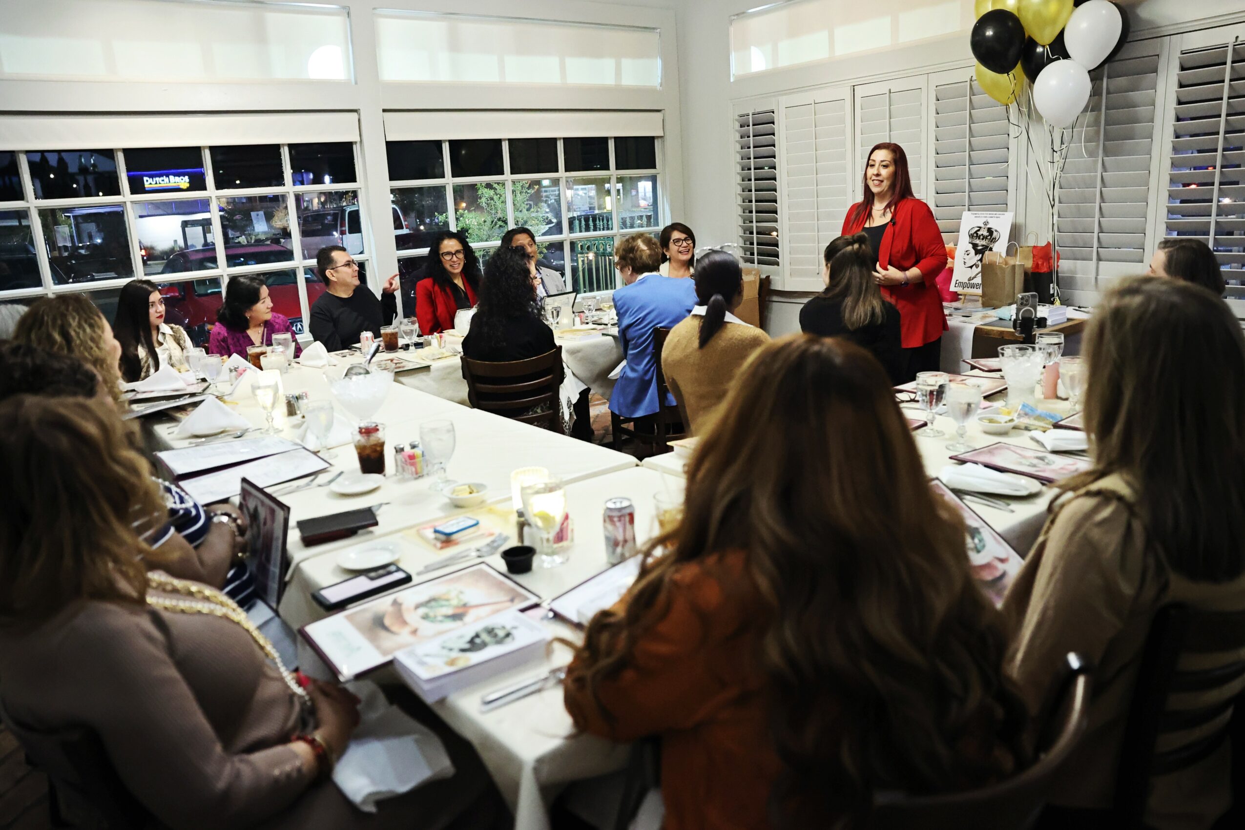 A woman in a red blazer stands at the head of a long table with people seated, in a room decorated with balloons.