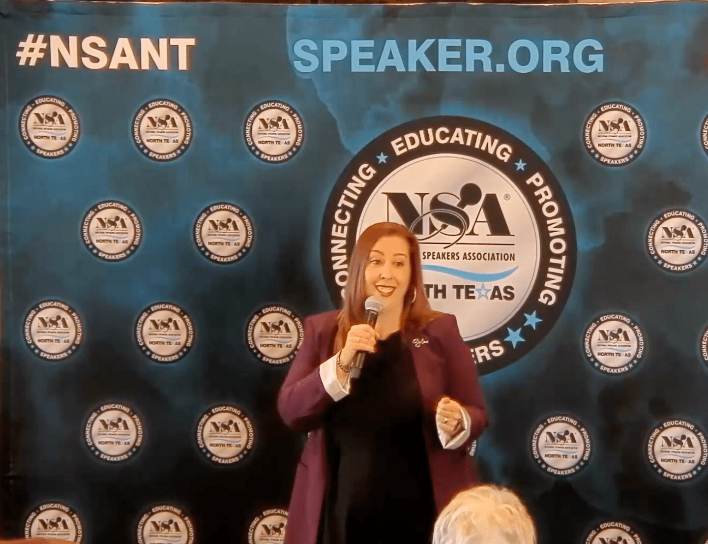 Woman speaking into microphone at event with backdrop displaying logos and text, including #NSANT, SPEAKER.ORG, and NSA. Confidence: 0.9, alt_text_word_count: 24