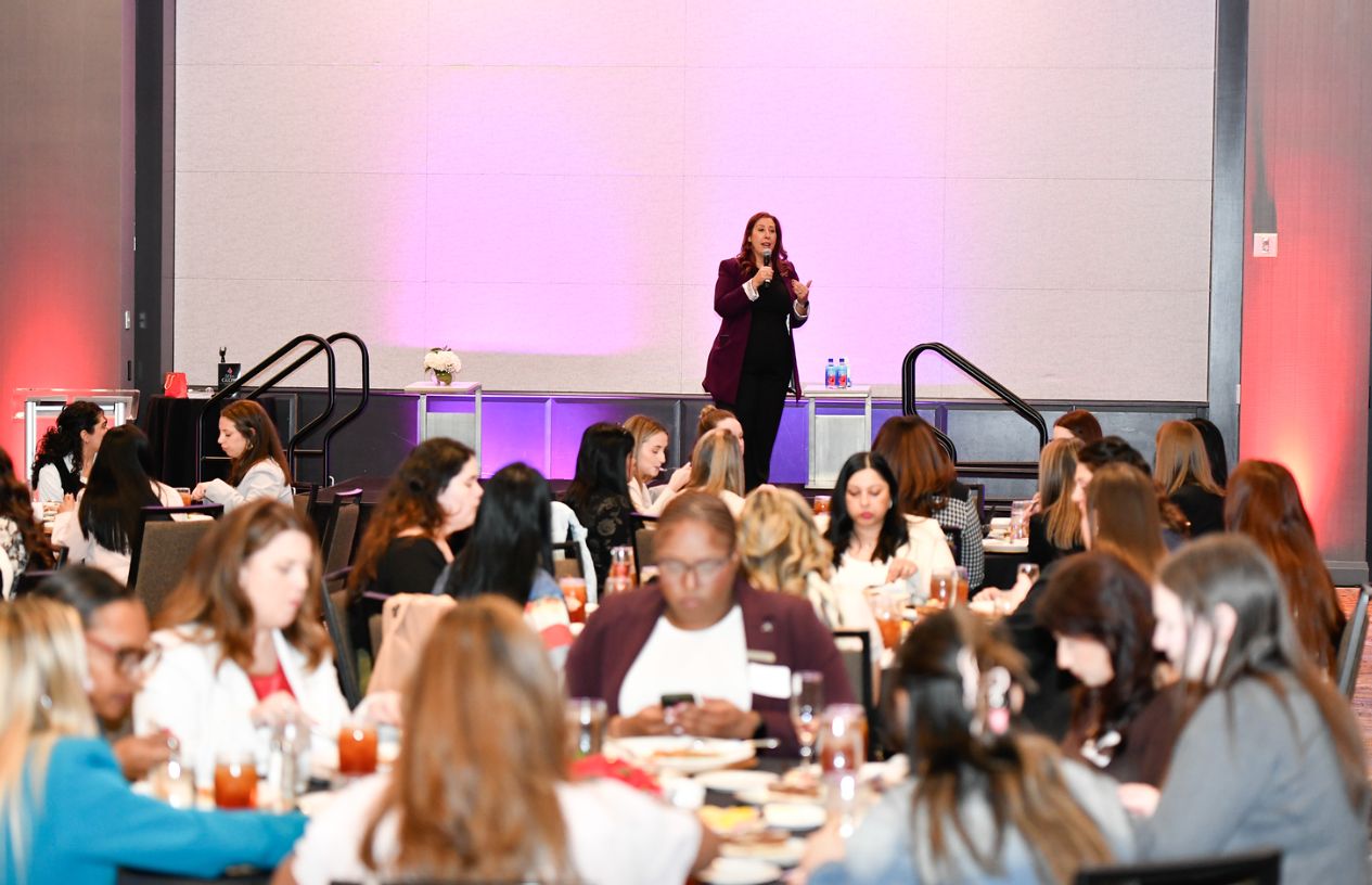 A woman stands on stage speaking to an audience in a large conference room with tables and attendees.