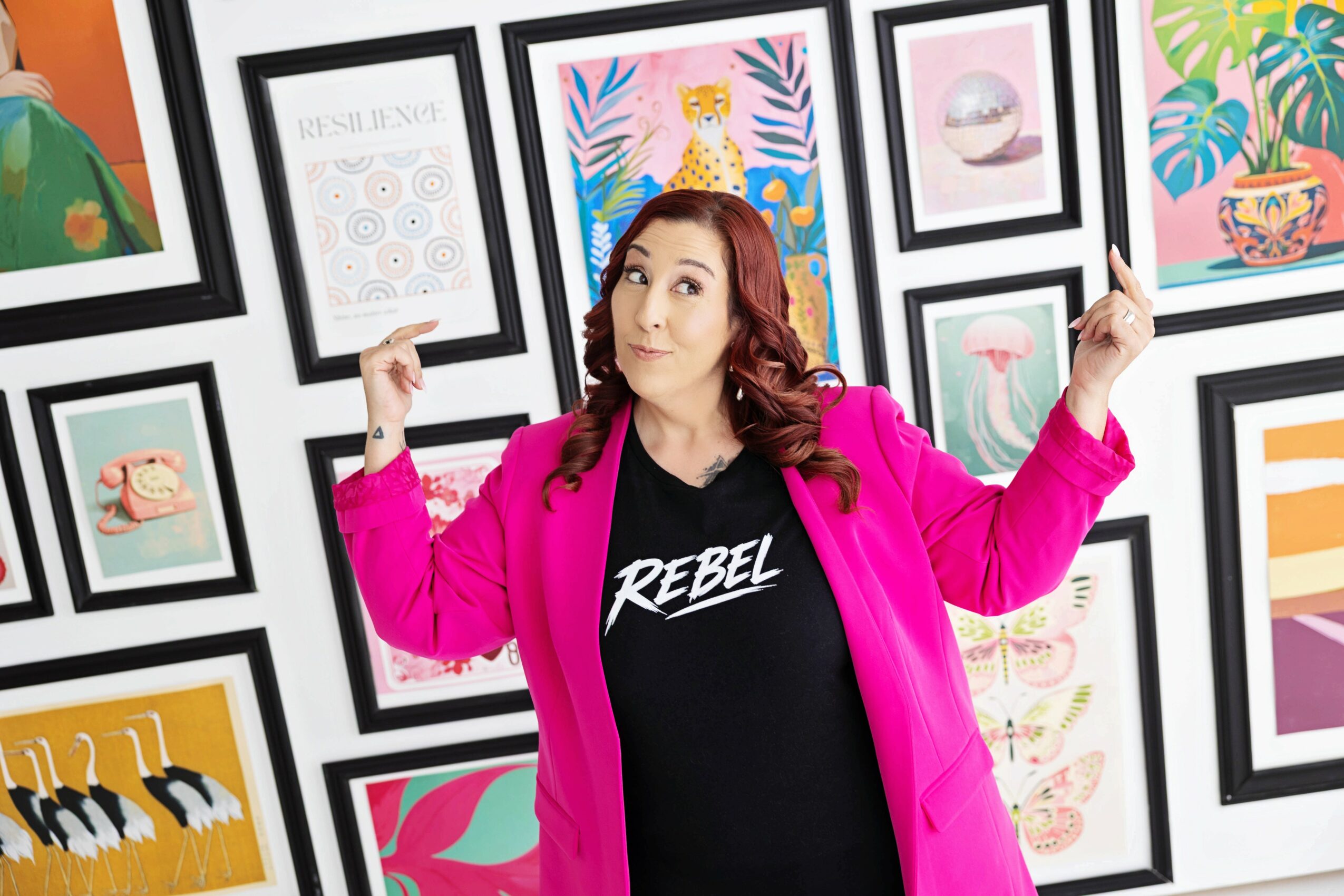 Woman with curly hair wearing a pink jacket and black shirt with 'REBEL' text, standing in front of a colorful framed art display, flexing her arms.