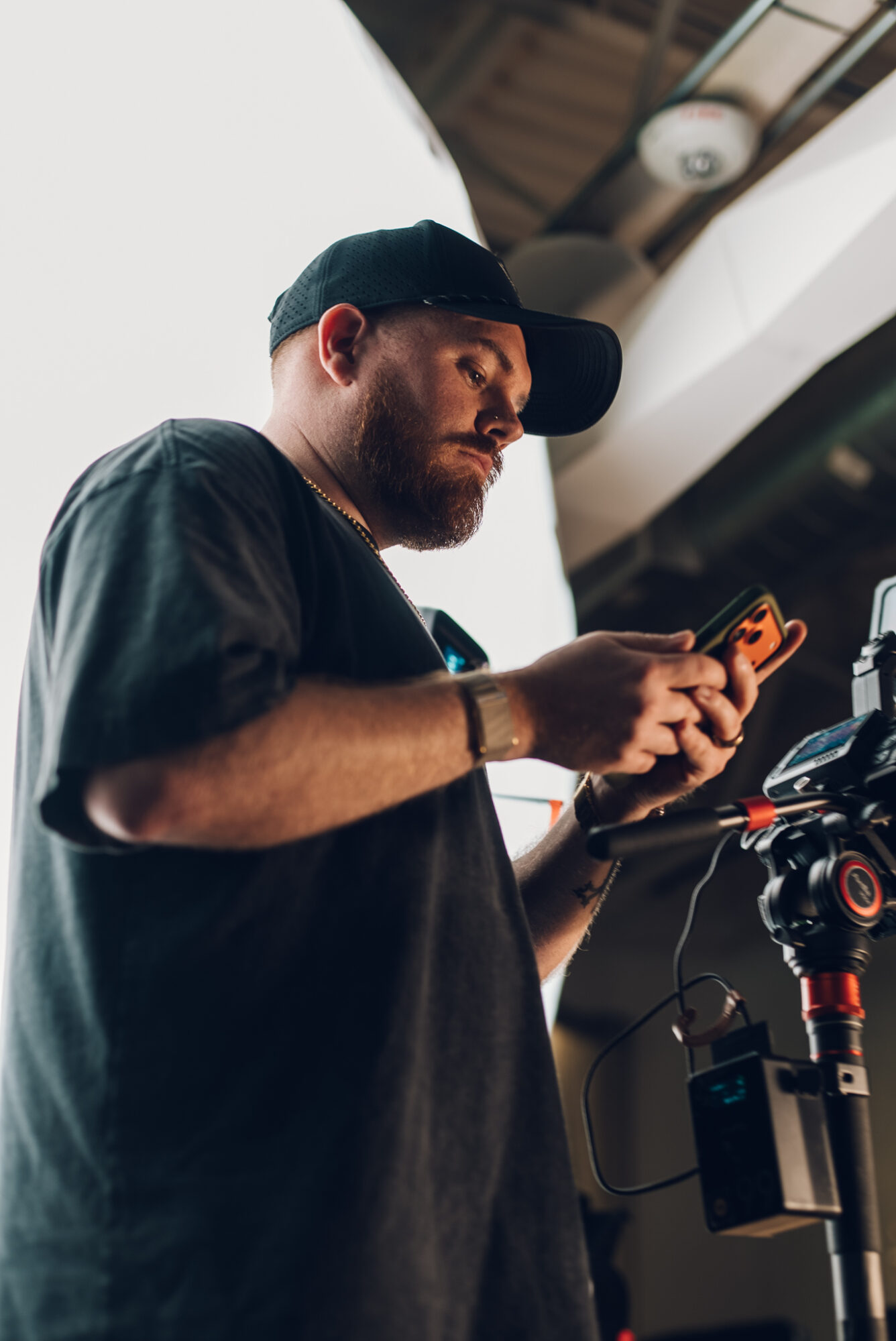 Man with a beard wearing a black cap and shirt, looking at a smartphone, with a camera mounted nearby.