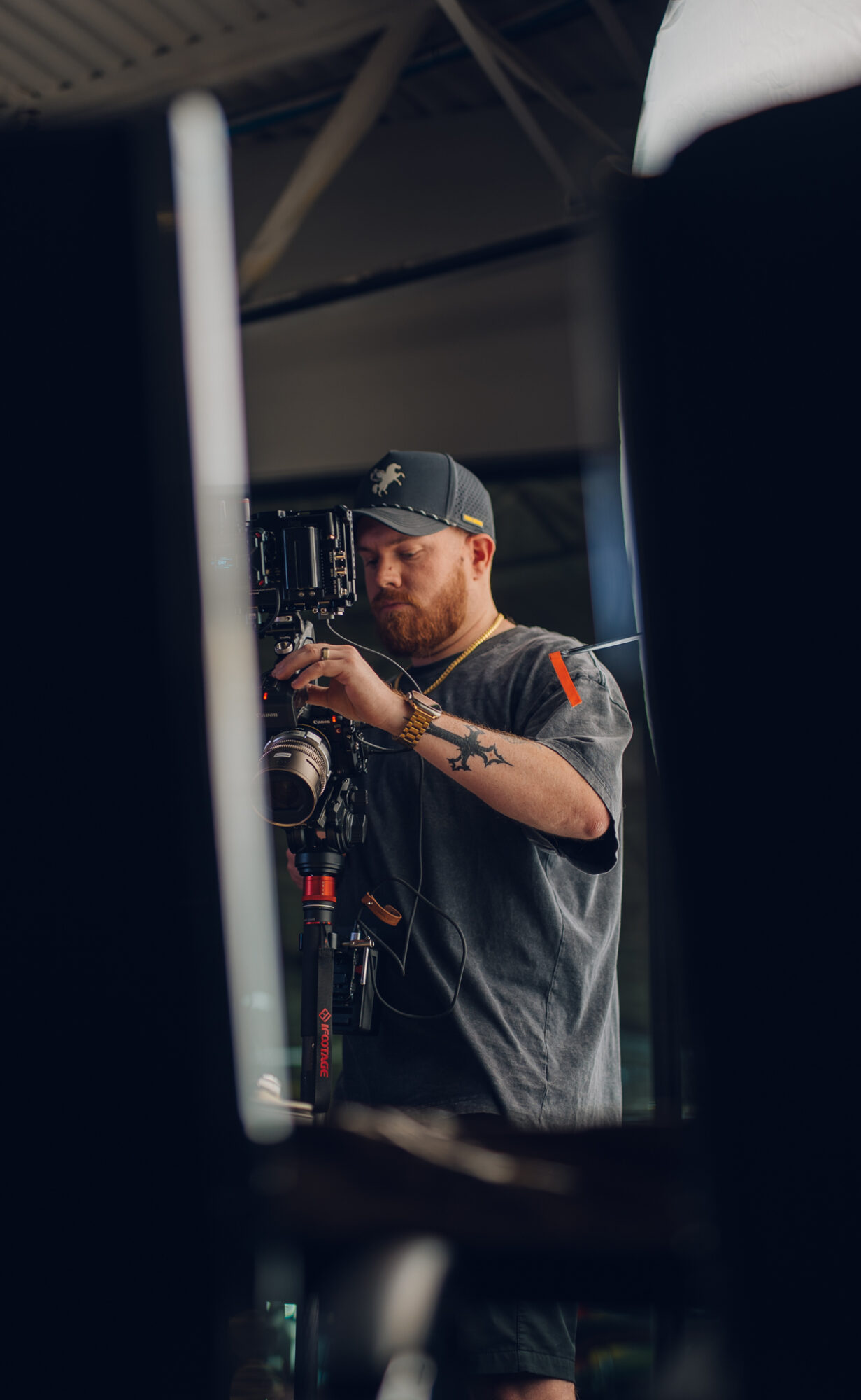 Person with a beard and cap adjusting camera equipment, reflected in a mirror or screen, in a dimly lit environment.