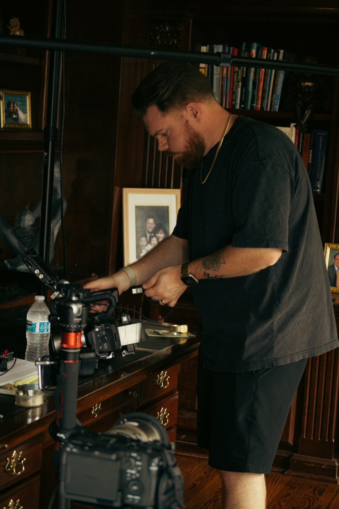 Man with beard and dark hair adjusting camera equipment in a room with bookshelves and framed photos