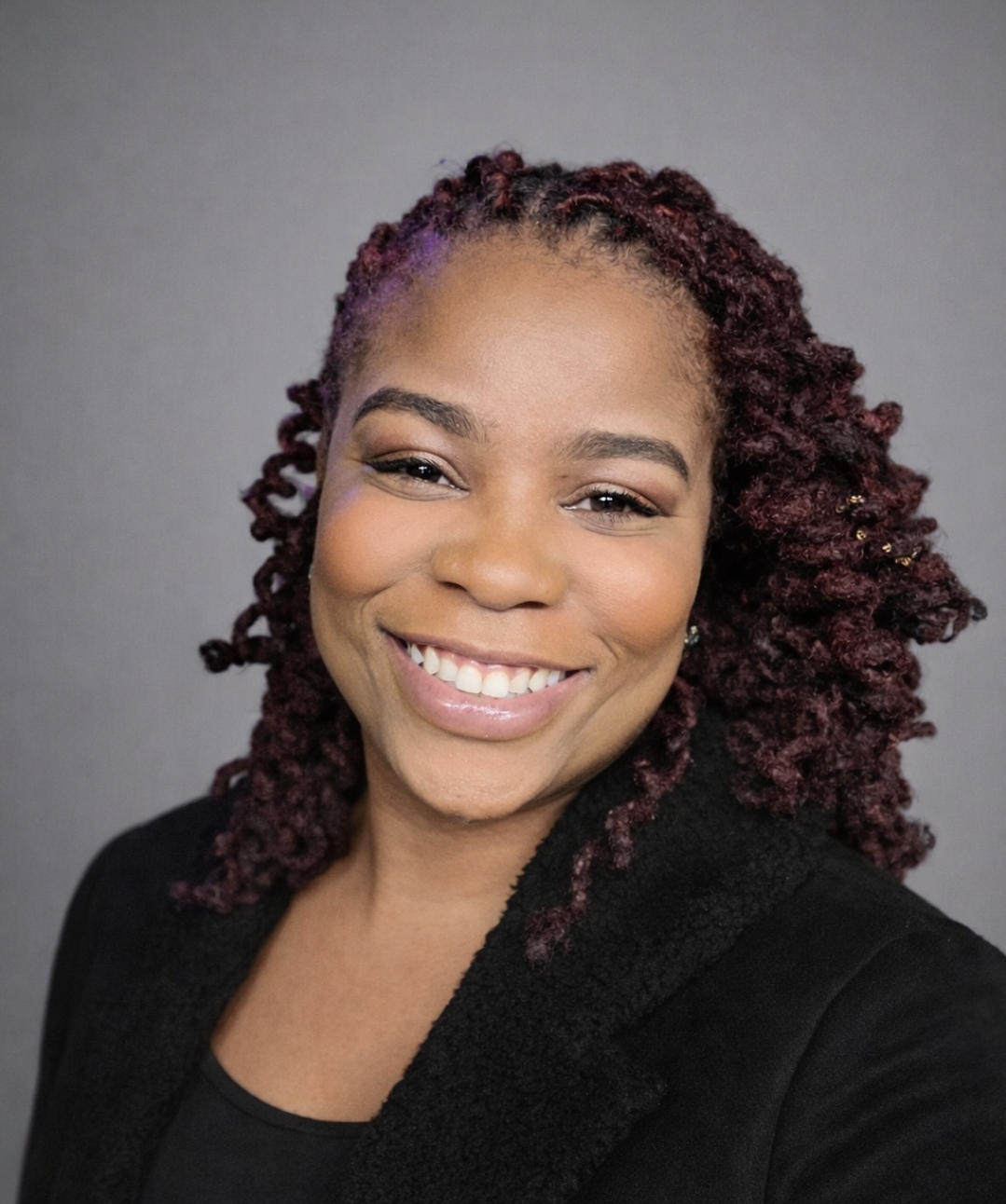Smiling woman with curly hair and a black top, facing forward, against a gray background.