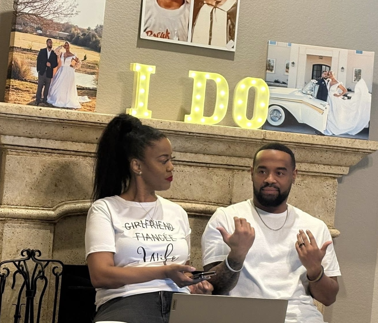 Two people sitting in front of a fireplace with wedding photos and illuminated letters spelling 'I DO'.
