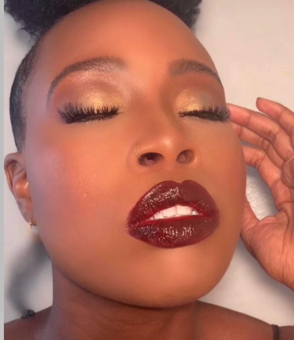 Close-up of a woman with closed eyes, gold eyeshadow, dark red lipstick, and hand near face.