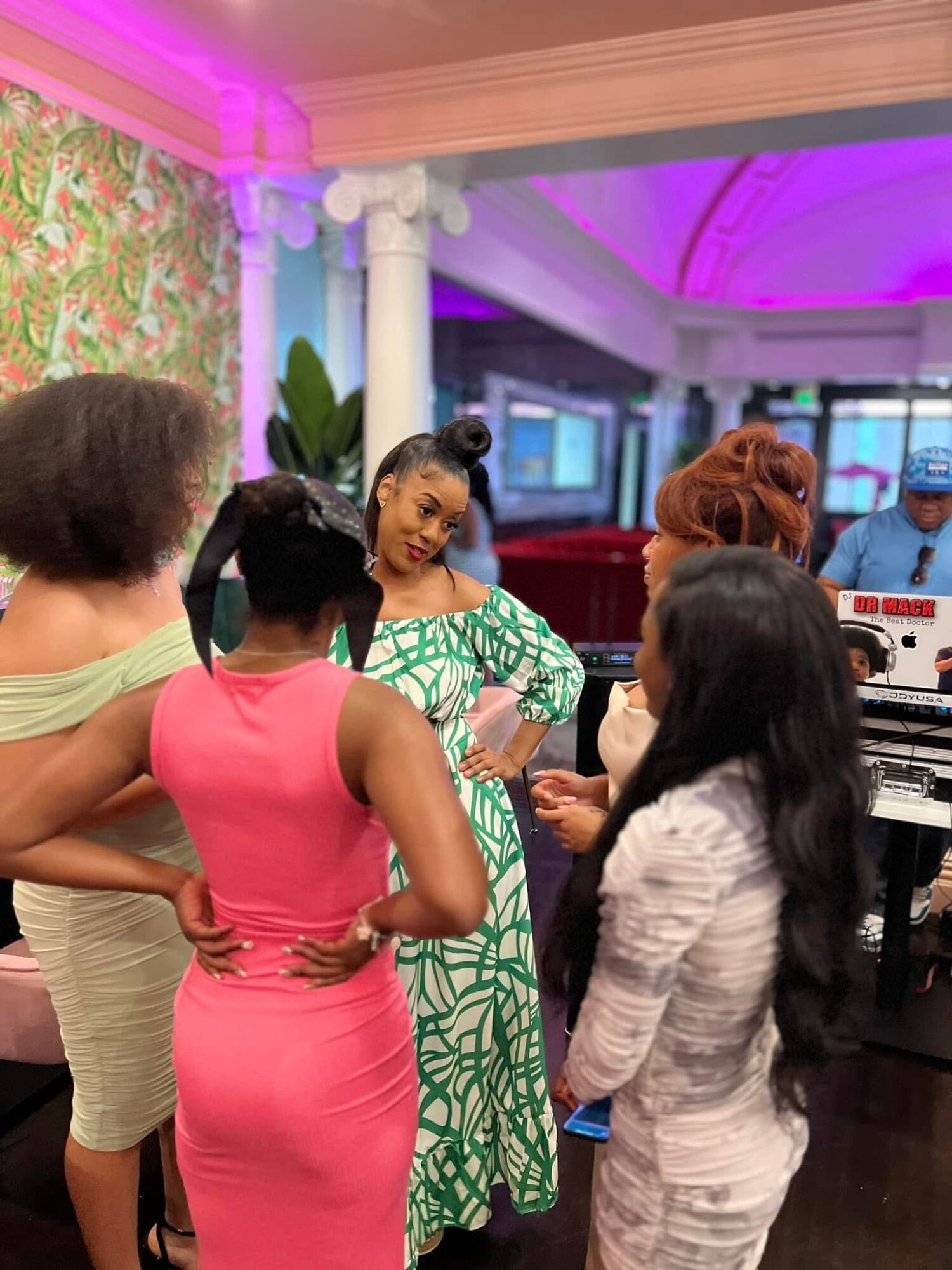 Group of women in conversation indoors with colorful lighting and decorative walls, some wearing patterned dresses.