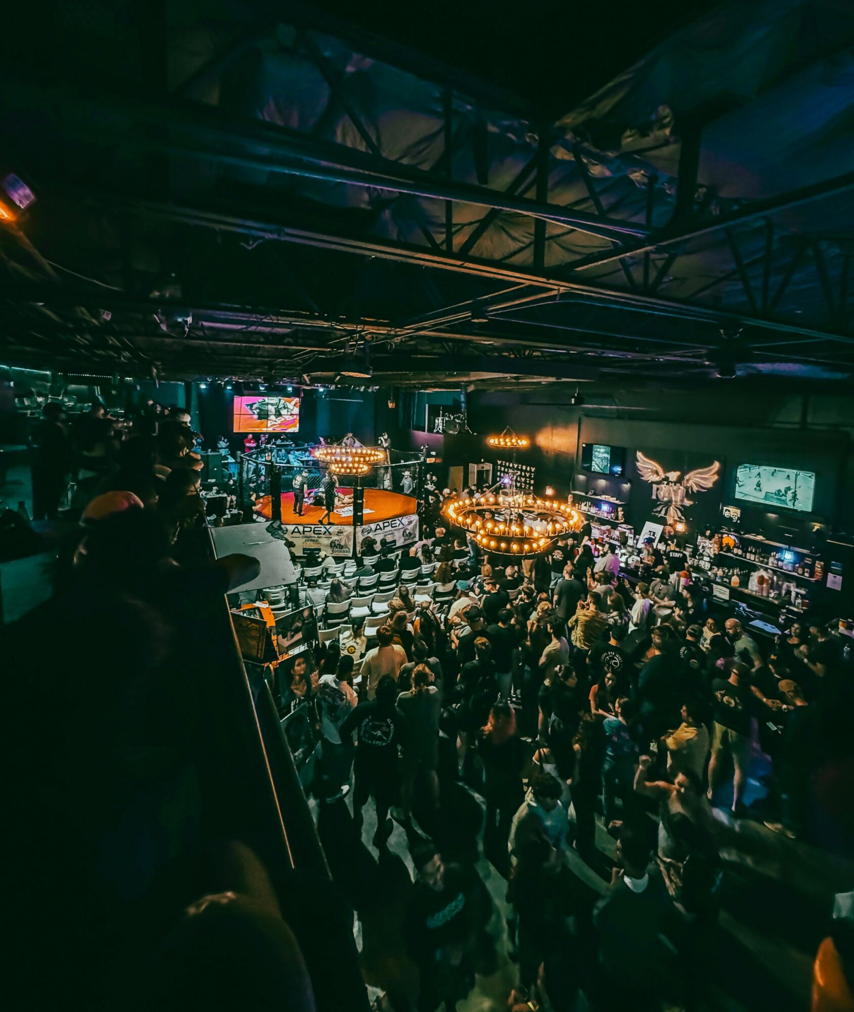 Crowd gathered in a dimly lit venue with stage, screens, and decorative lighting, viewed from an elevated angle.