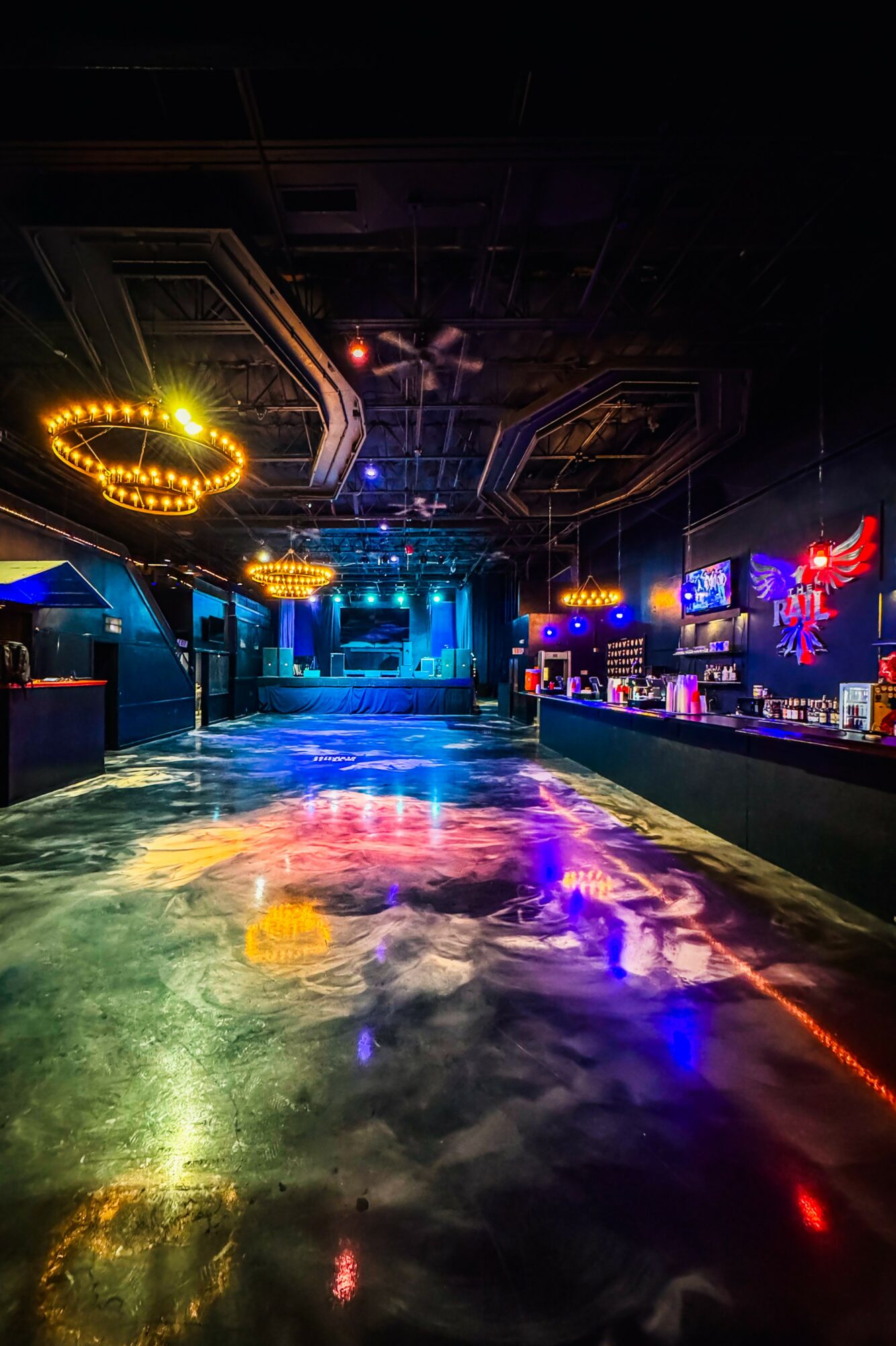 Empty nightclub with colorful lighting, illuminated bar, and a reflective dance floor, with a dark ceiling and neon signs.