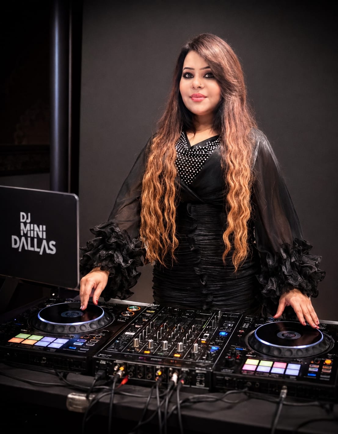 Woman with long wavy hair standing behind DJ equipment, wearing a black dress with ruffled sleeves, smiling.