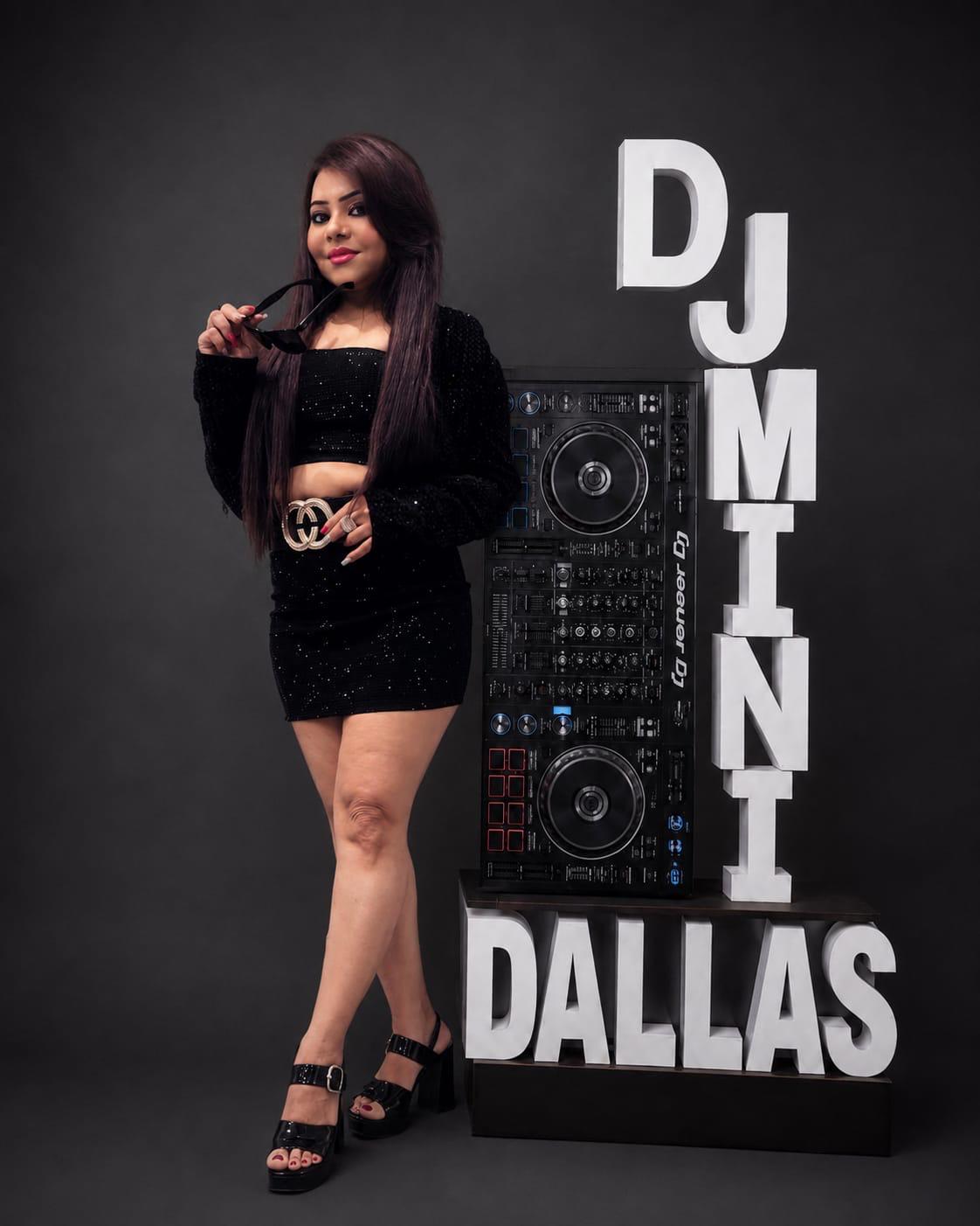 Woman with long dark hair in black outfit standing next to DJ equipment and large white letters spelling DUMINI DALLAS.