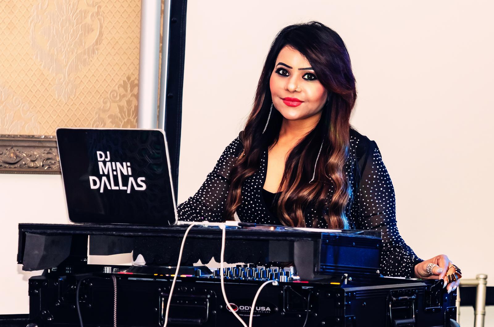 Woman DJ with long dark hair, wearing a black polka dot top, standing behind DJ equipment, smiling at the camera.