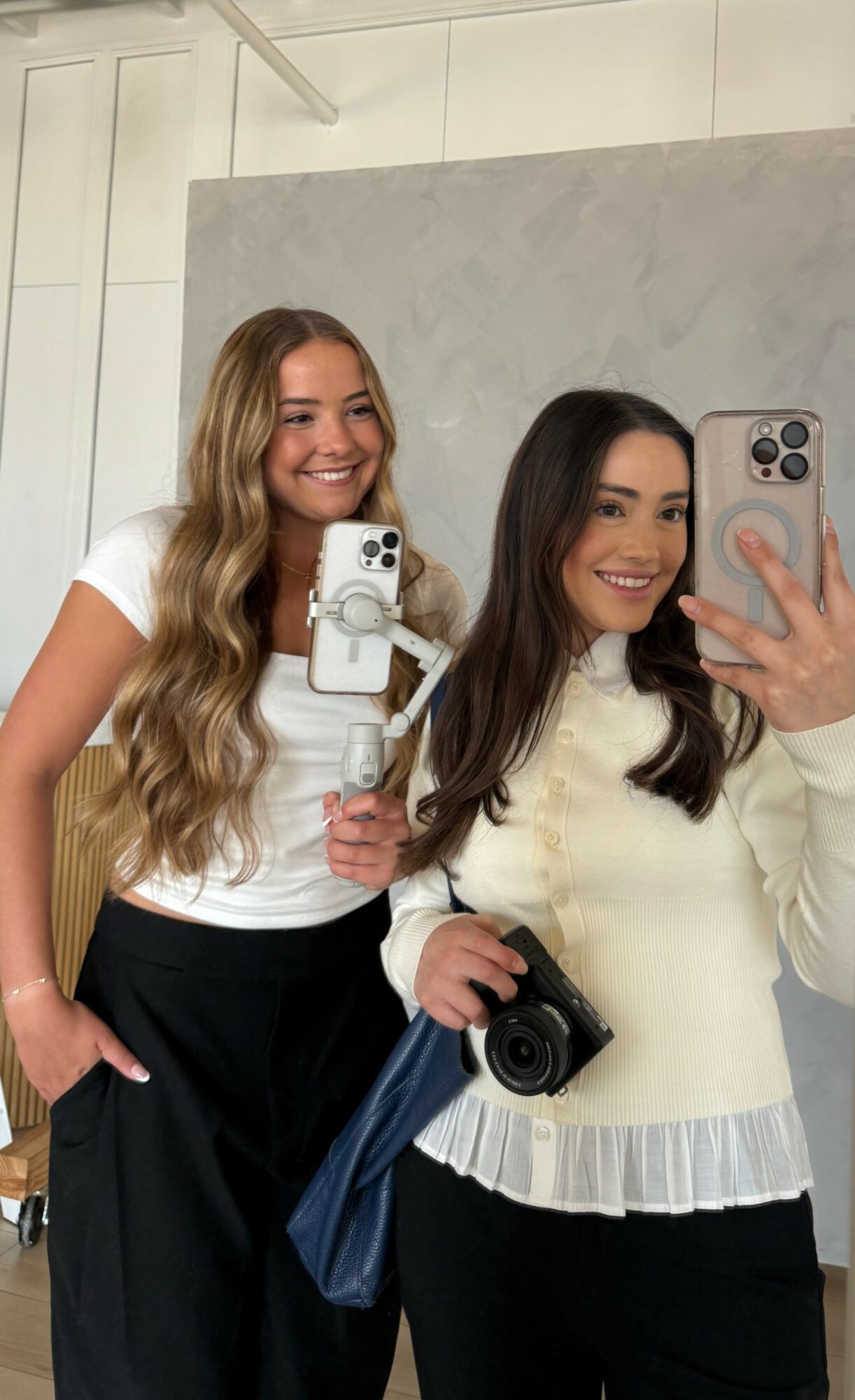 Two women smiling, taking a mirror selfie with a camera, one holding a phone and the other a camera, in a room with a gray wall.