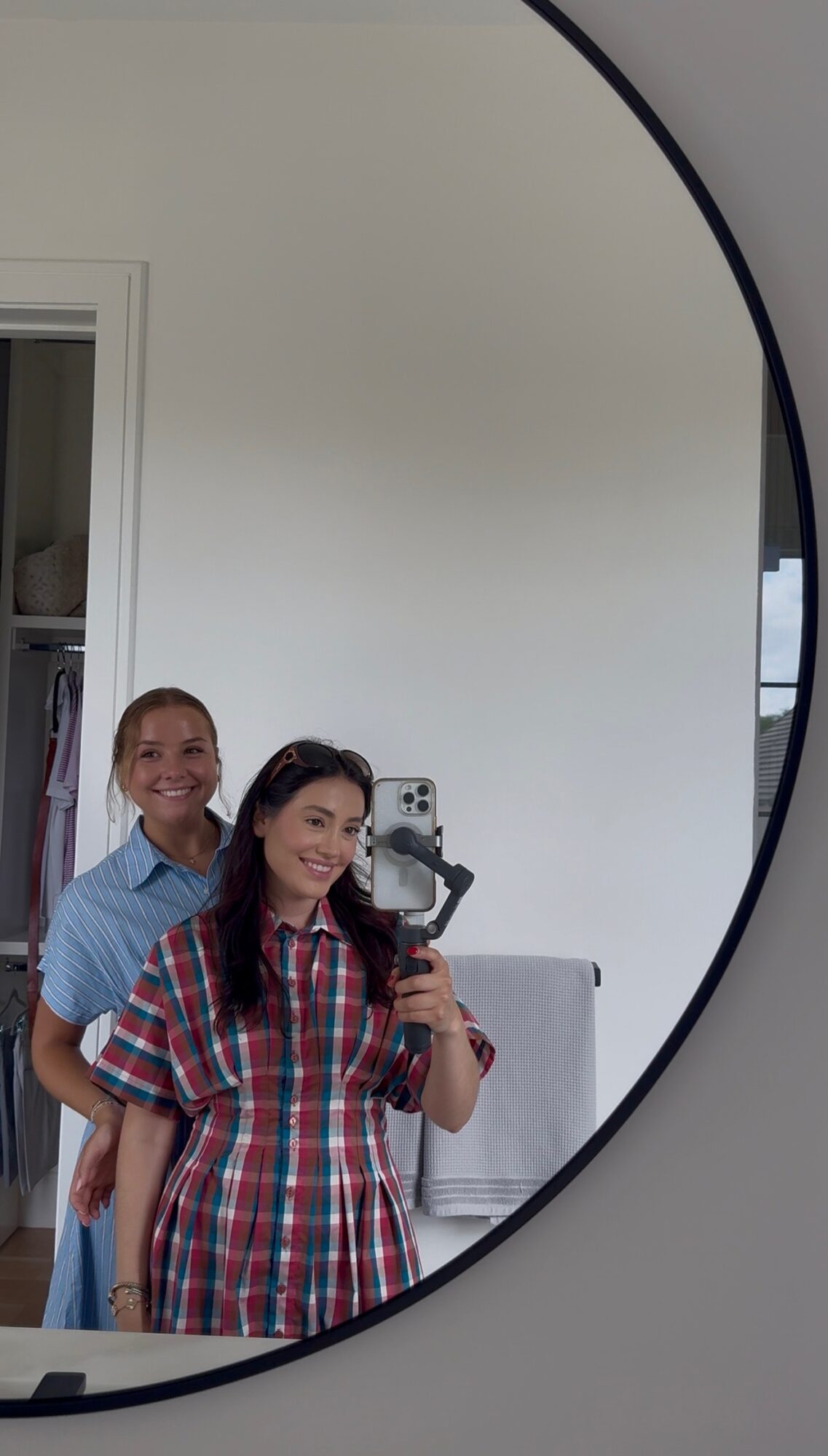Two women taking a mirror selfie, one holding a phone, smiling, in a room with a white wall and door.