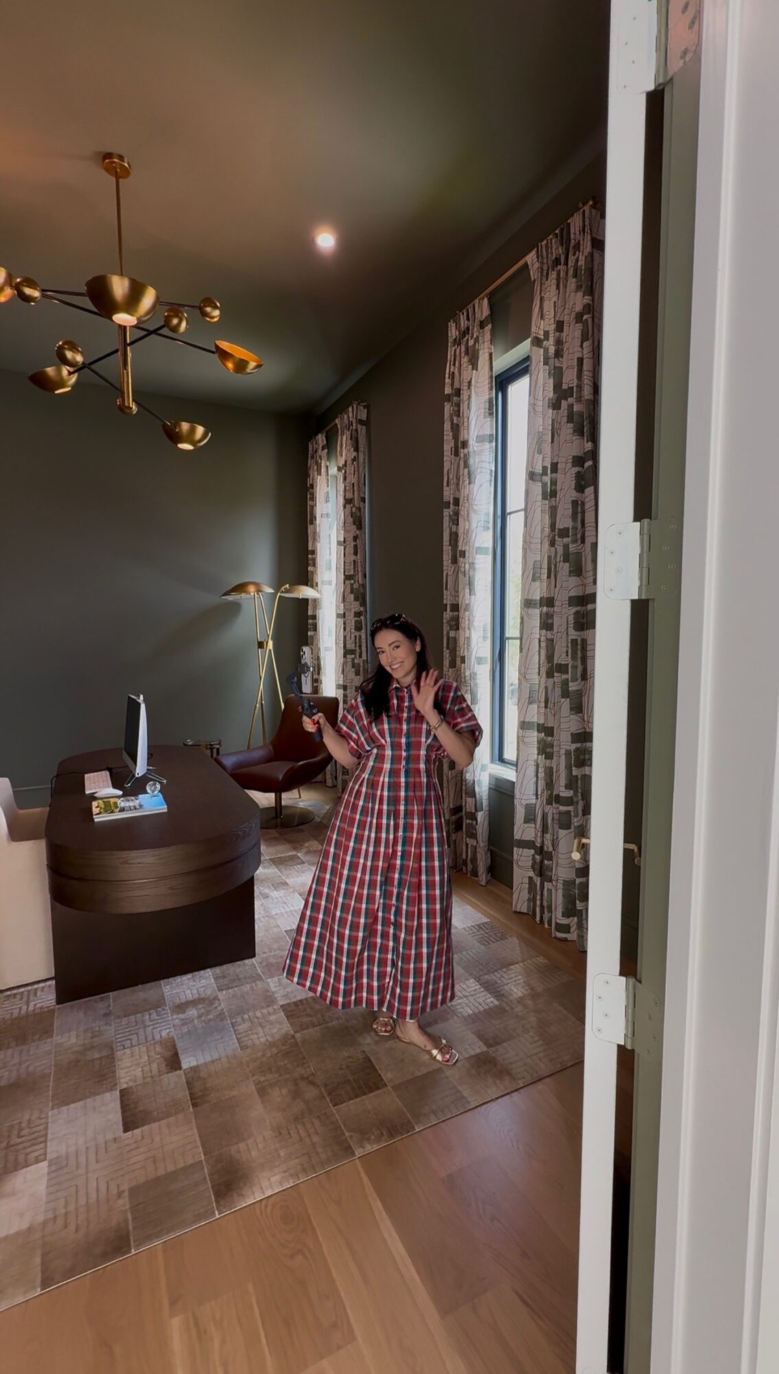 Person in a long checkered dress standing in a room with tall windows and patterned curtains, holding a phone, with a chandelier overhead.