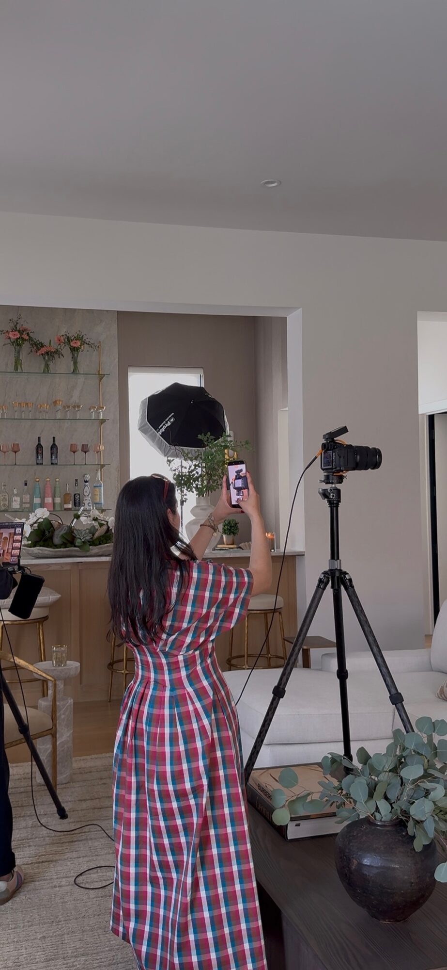 Woman in plaid dress takes photo with smartphone in a room with camera equipment and decor.