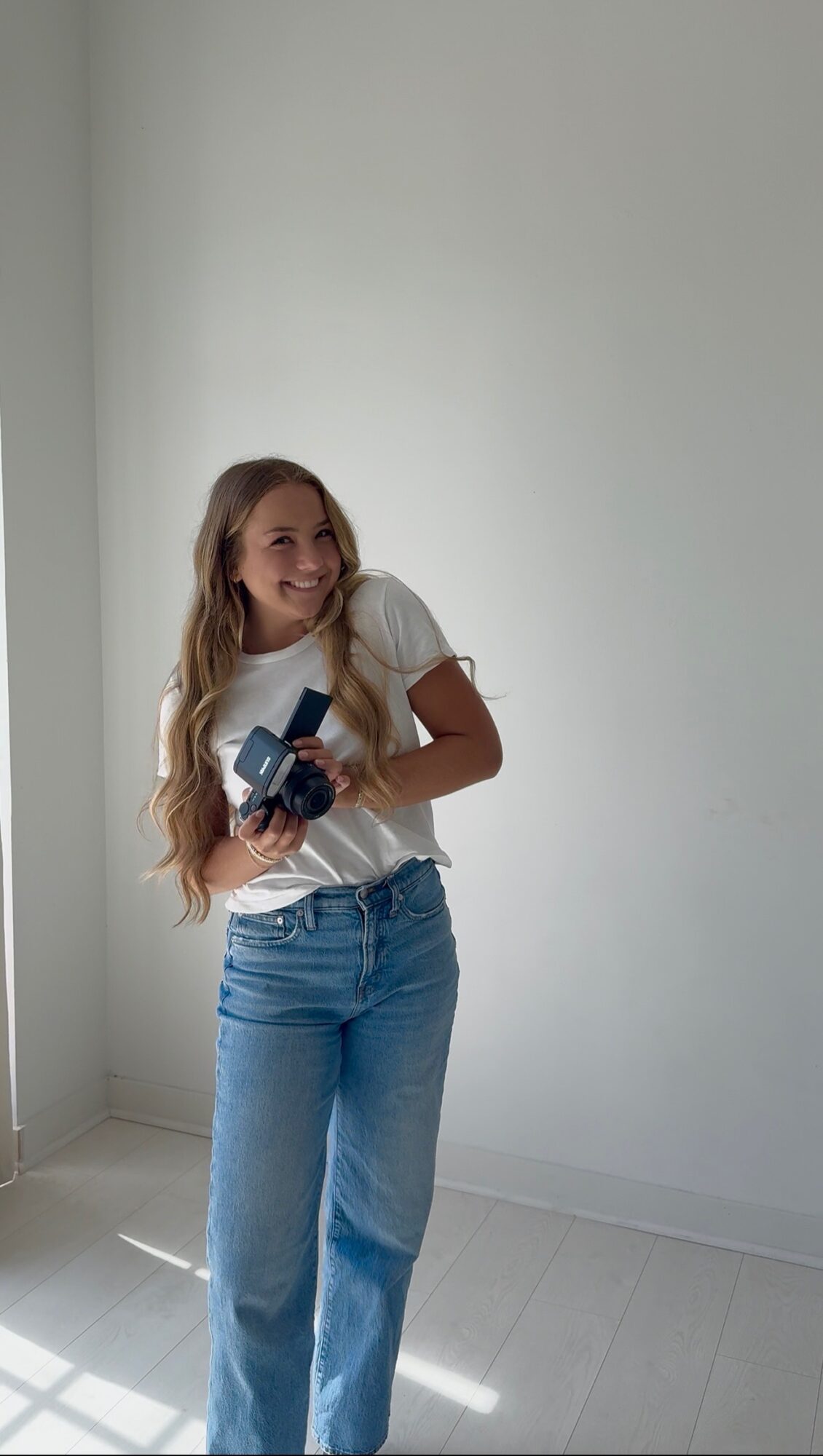 Young woman with long hair holding a camera, smiling, standing in a bright room with white walls and floor.