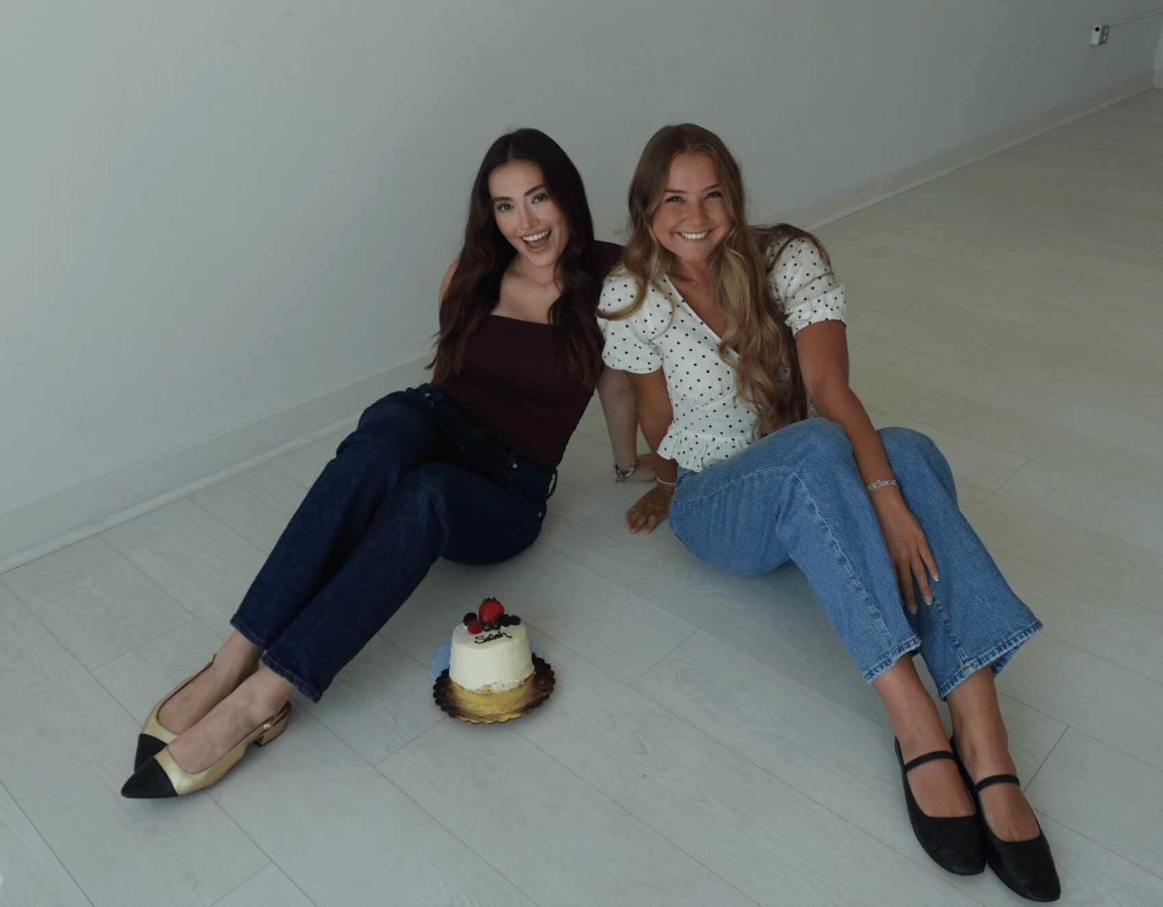 Two women sitting on the floor with a small cake between them, smiling, against a plain wall and light-colored floor.