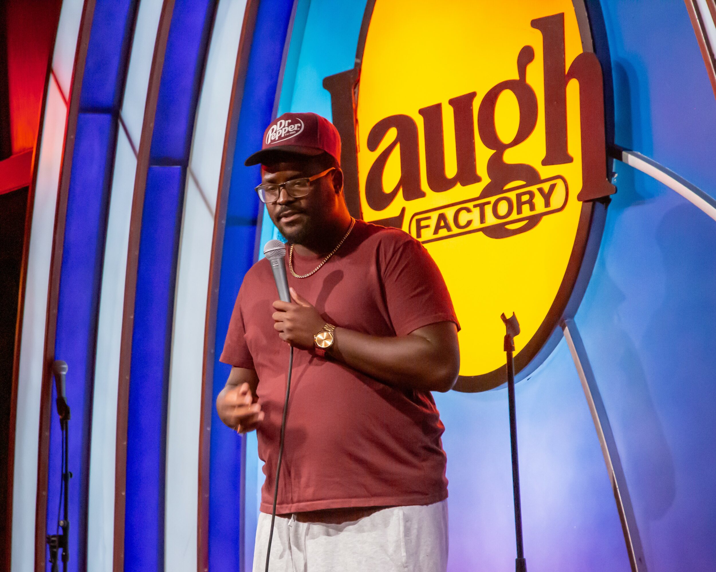 Person standing on stage in front of Laugh Factory sign, wearing a maroon shirt, cap, and glasses, holding a microphone.