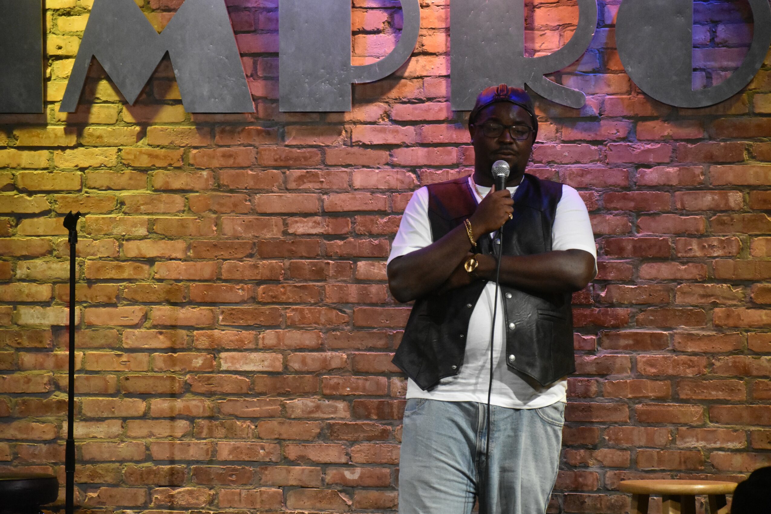 Person speaking into microphone on stage with brick wall background, large letters partially visible, wearing glasses, vest, and white shirt.